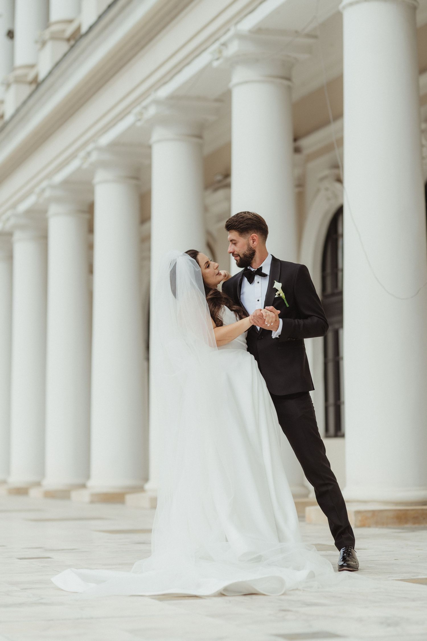 Romantic newlyweds sharing an intimate moment while holding hands among white classical columns.