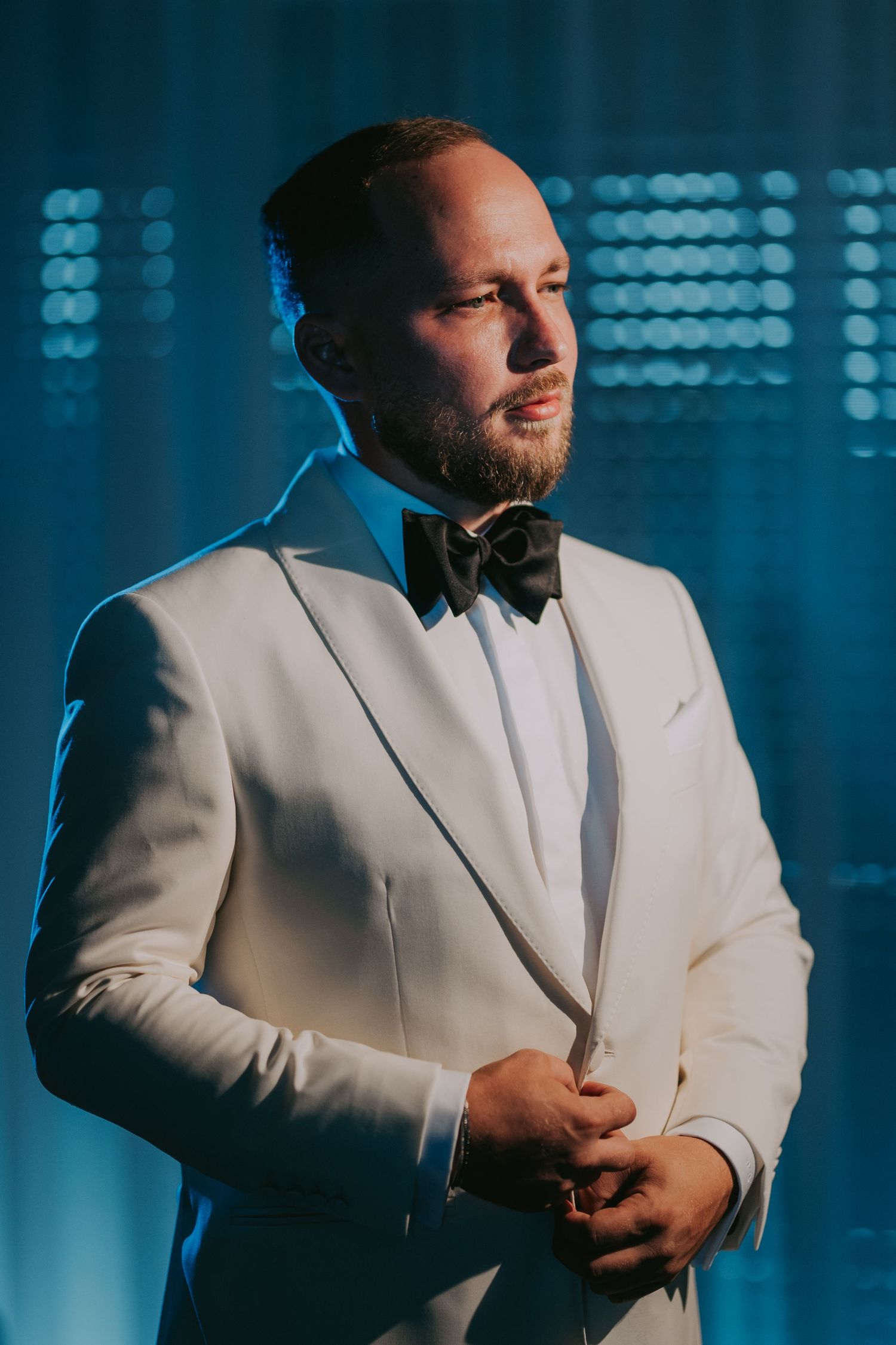 Dapper groom in white tuxedo and black bow tie posing with blue bokeh background. Epicu Fotograf Drobeta-Turnu Severin