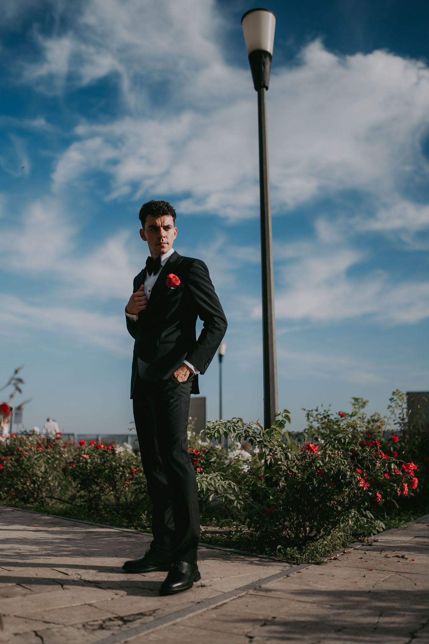 Stylish groom in black suit with red boutonniere posing by a lamppost surrounded by rose bushes. Epicu Fotograf Drobeta-Turnu Severin