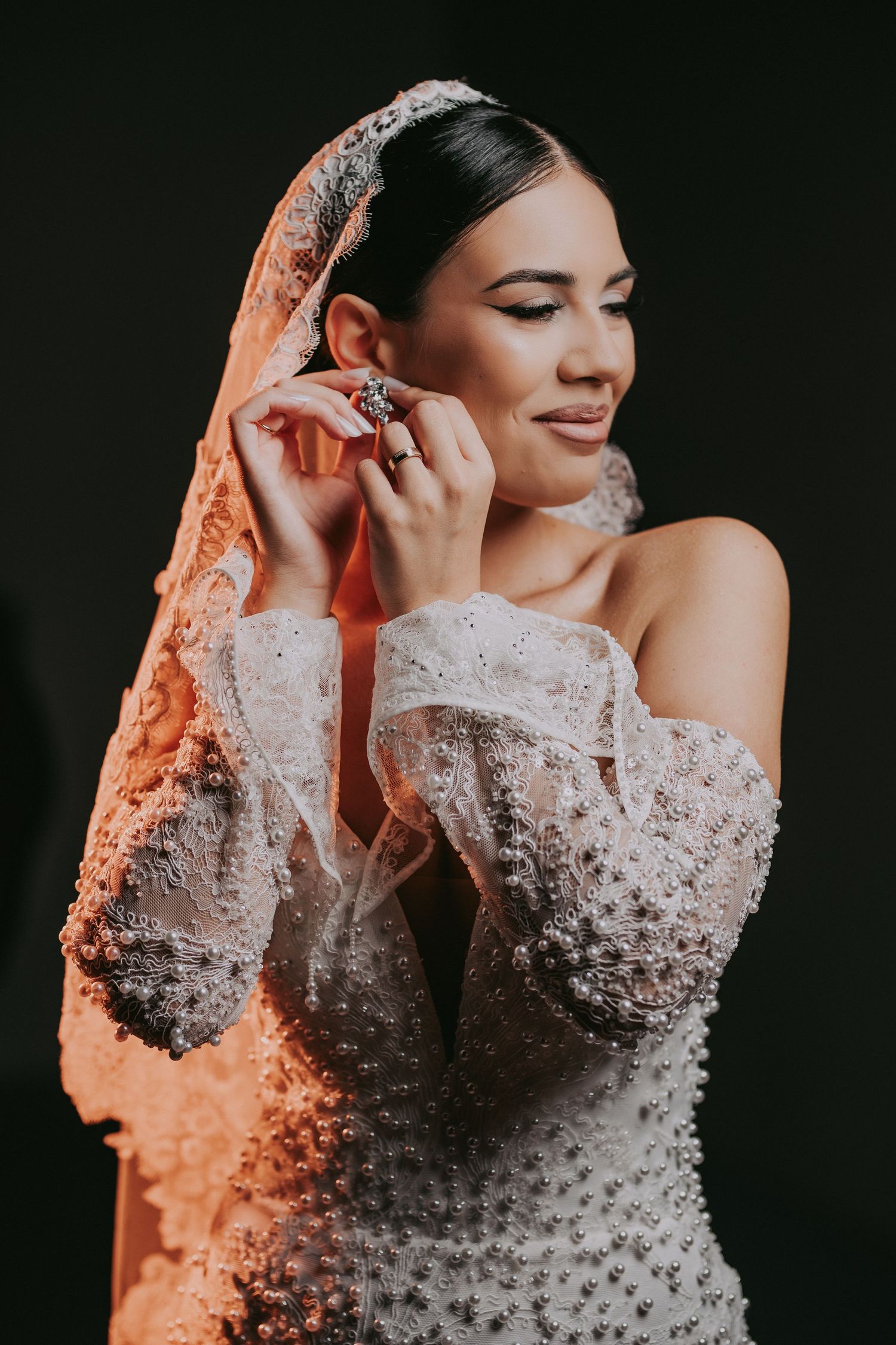 Bride in pearl-studded wedding gown adjusts earring with lace veil draped over head.