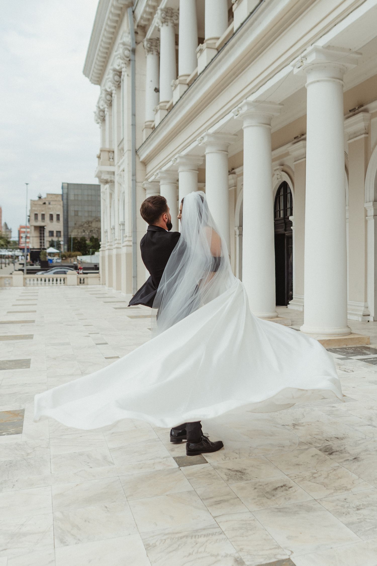 Bride's veil flowing dramatically in the wind as couple walks along a marble columned terrace.