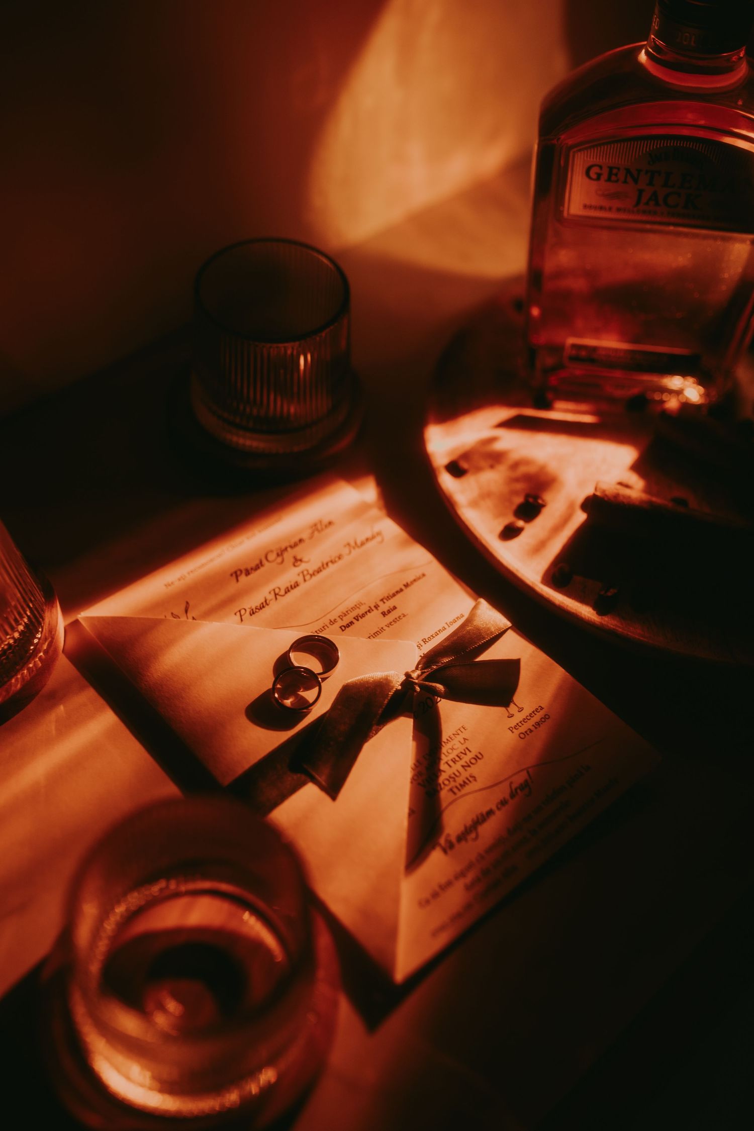 Wedding rings on invitation card beside Gentleman Jack whiskey bottle in warm red light.