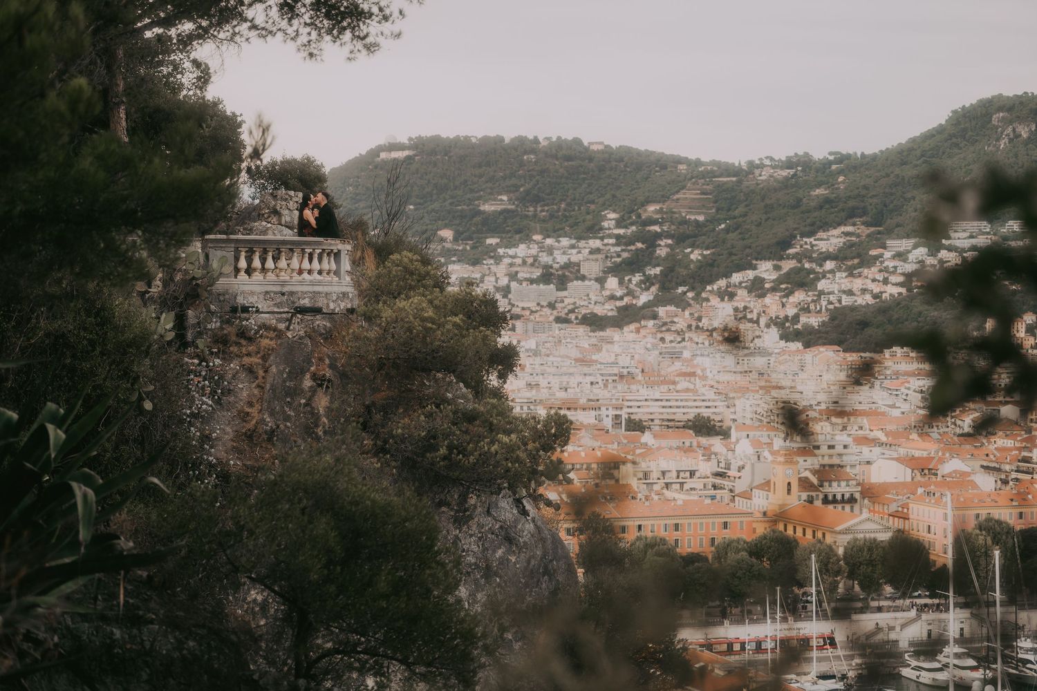 Couple stands on scenic overlook above picturesque coastal town with harbor and lush hillside views.