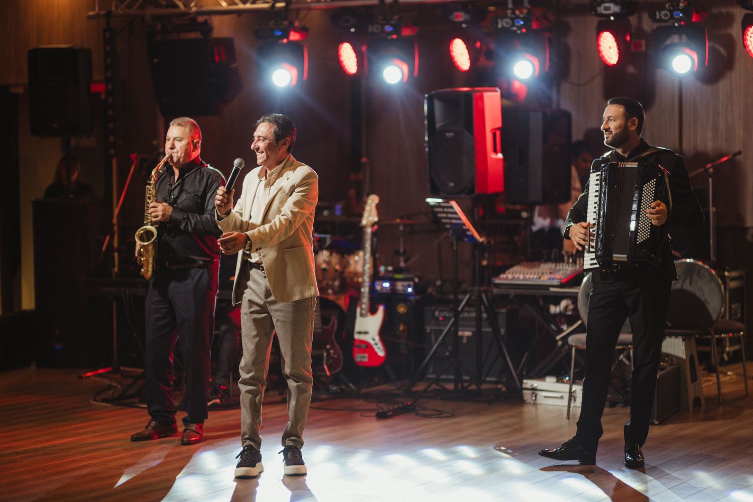Three male musicians performing on stage with saxophone, microphone, and accordion.