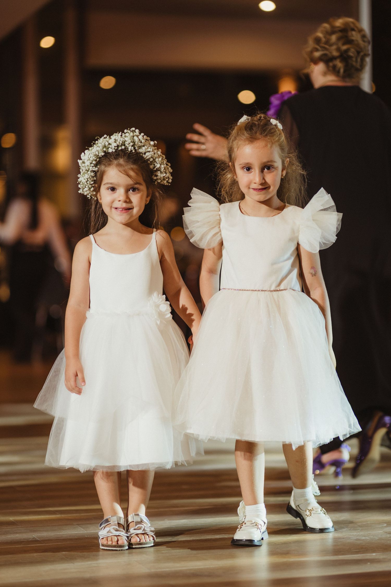 Two little flower girls in white tulle dresses holding hands at a wedding reception.