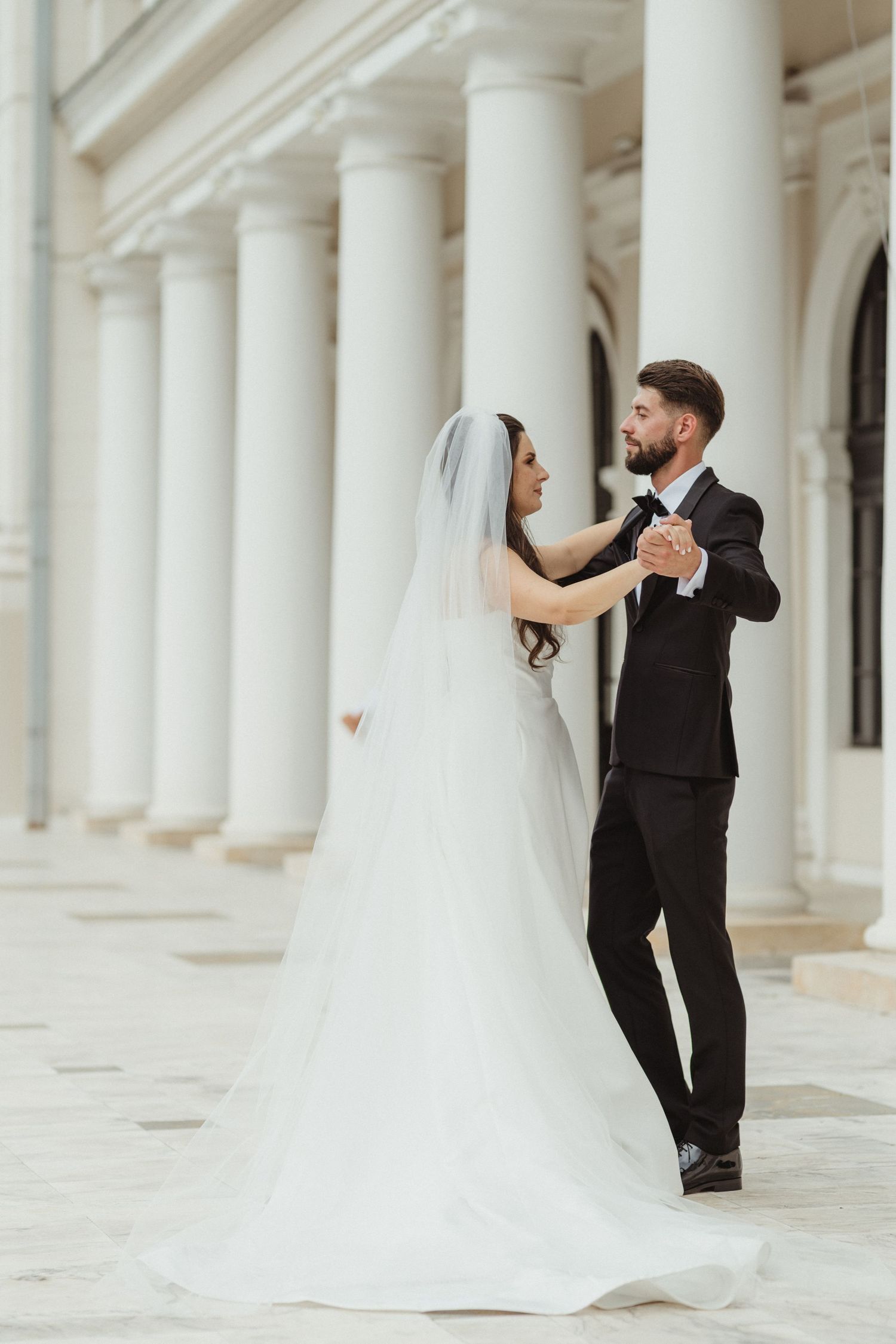 Elegant bride and groom dancing together in a grand white columned hall on their wedding day.