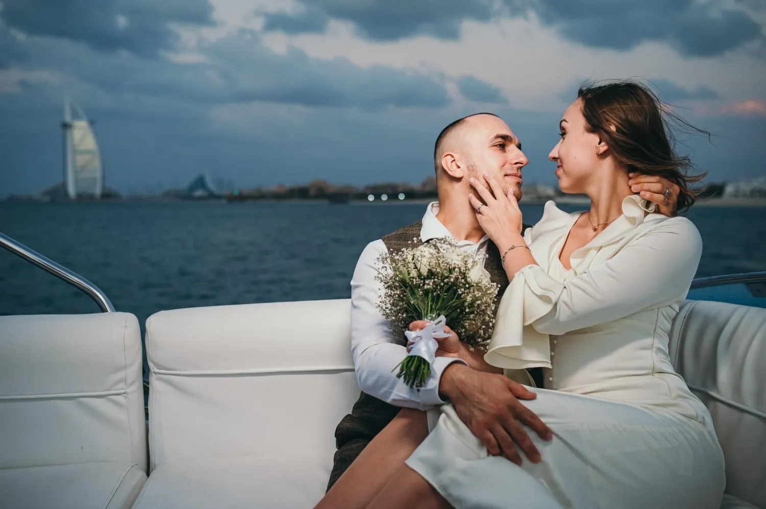 Bride and groom sitting on white yacht with bouquet, Burj Al Arab visible in background.