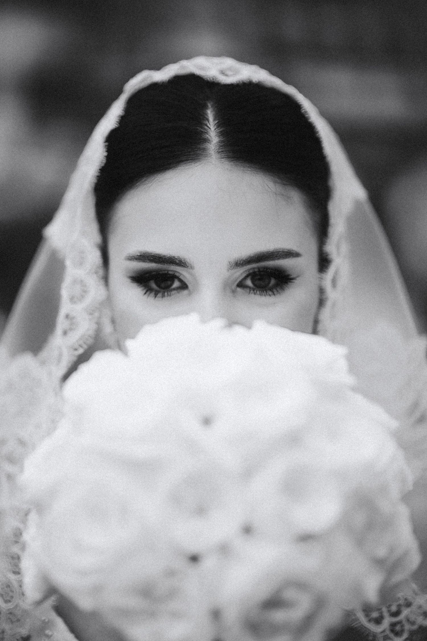 Black and white portrait of bride with lace veil holding white rose bouquet over face.