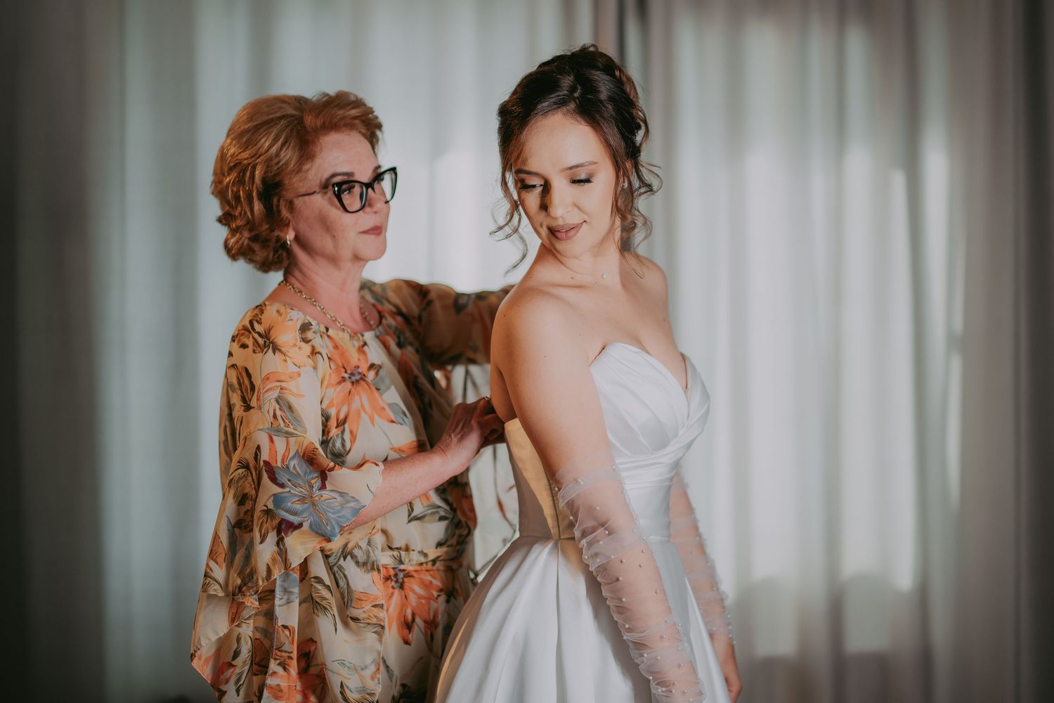 Mother helping bride zip up her elegant white wedding gown near a bright window. Epicu Fotograf Drobeta-Turnu Severin