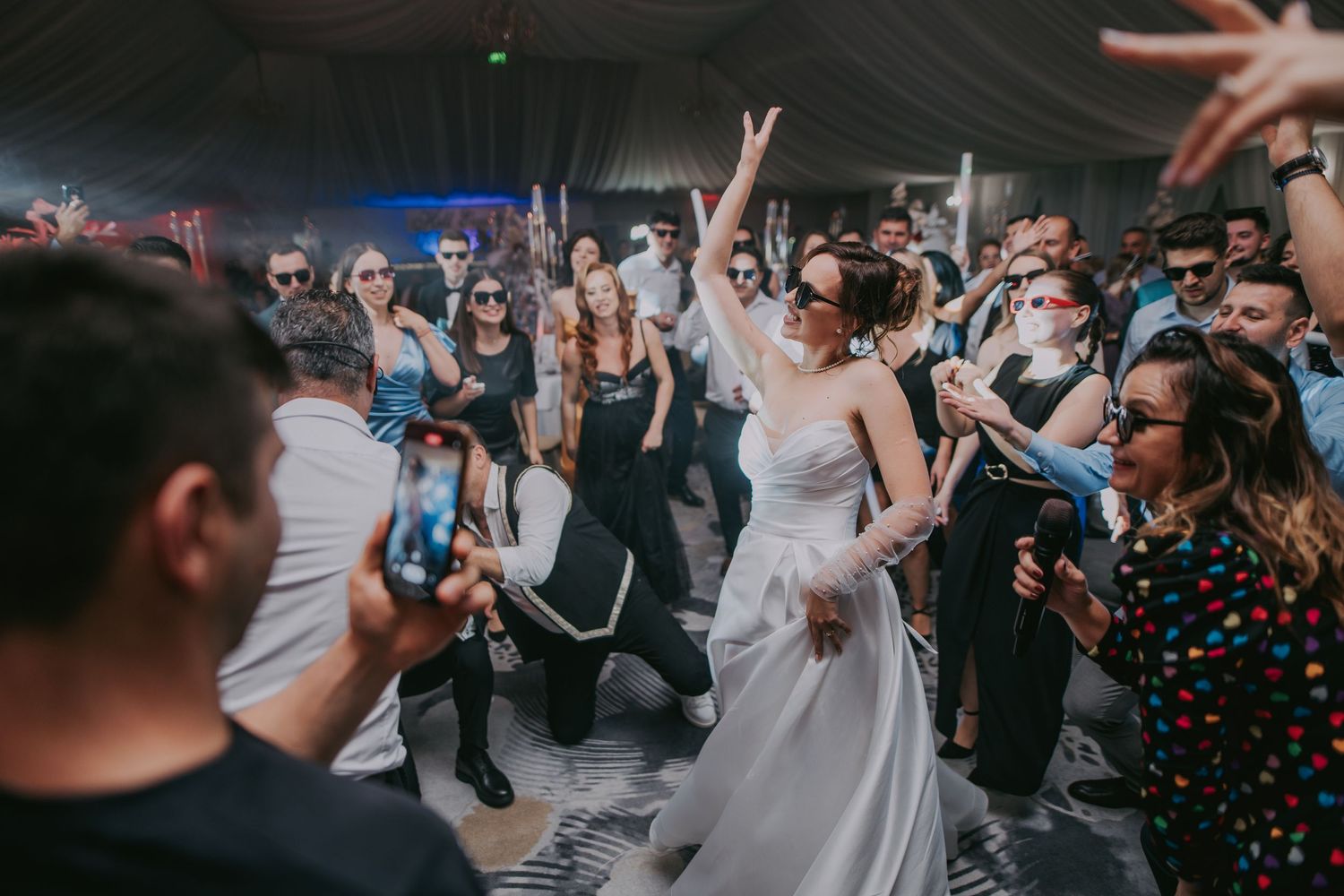 Bride in white dress dancing with one arm raised among celebrating wedding guests at indoor reception. Epicu Fotograf Drobeta-Turnu Severin