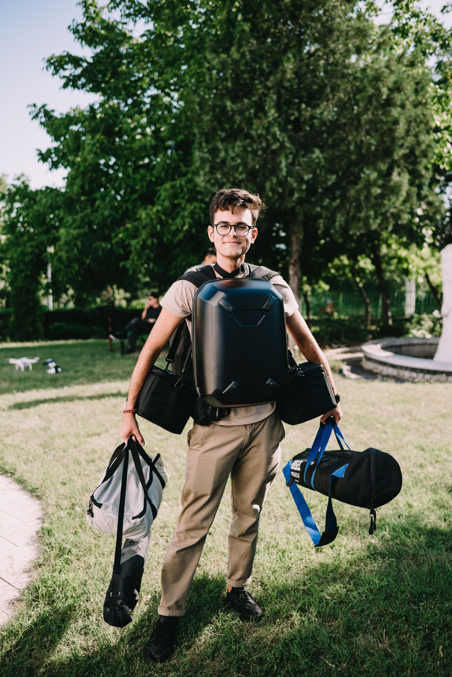 Young man with glasses carrying multiple bags and a hard shell backpack in a sunny green park.