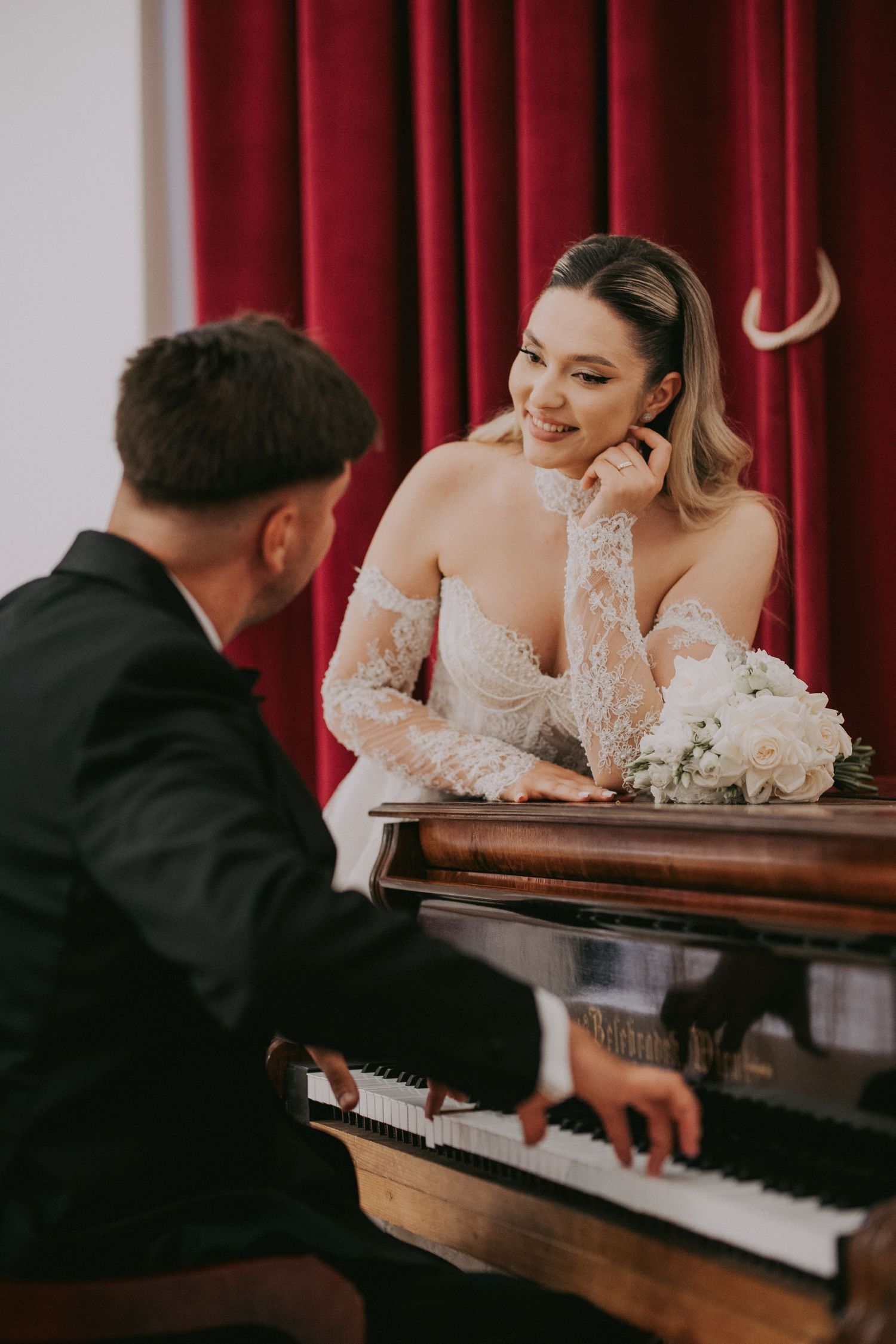 Smiling bride in lace gown leans on grand piano as groom plays at elegant wedding.