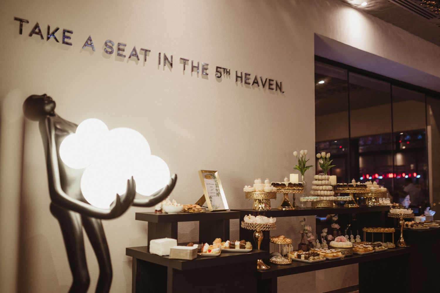 Stylish dessert display table at upscale venue with Take a Seat in the 5th Heaven wall quote.