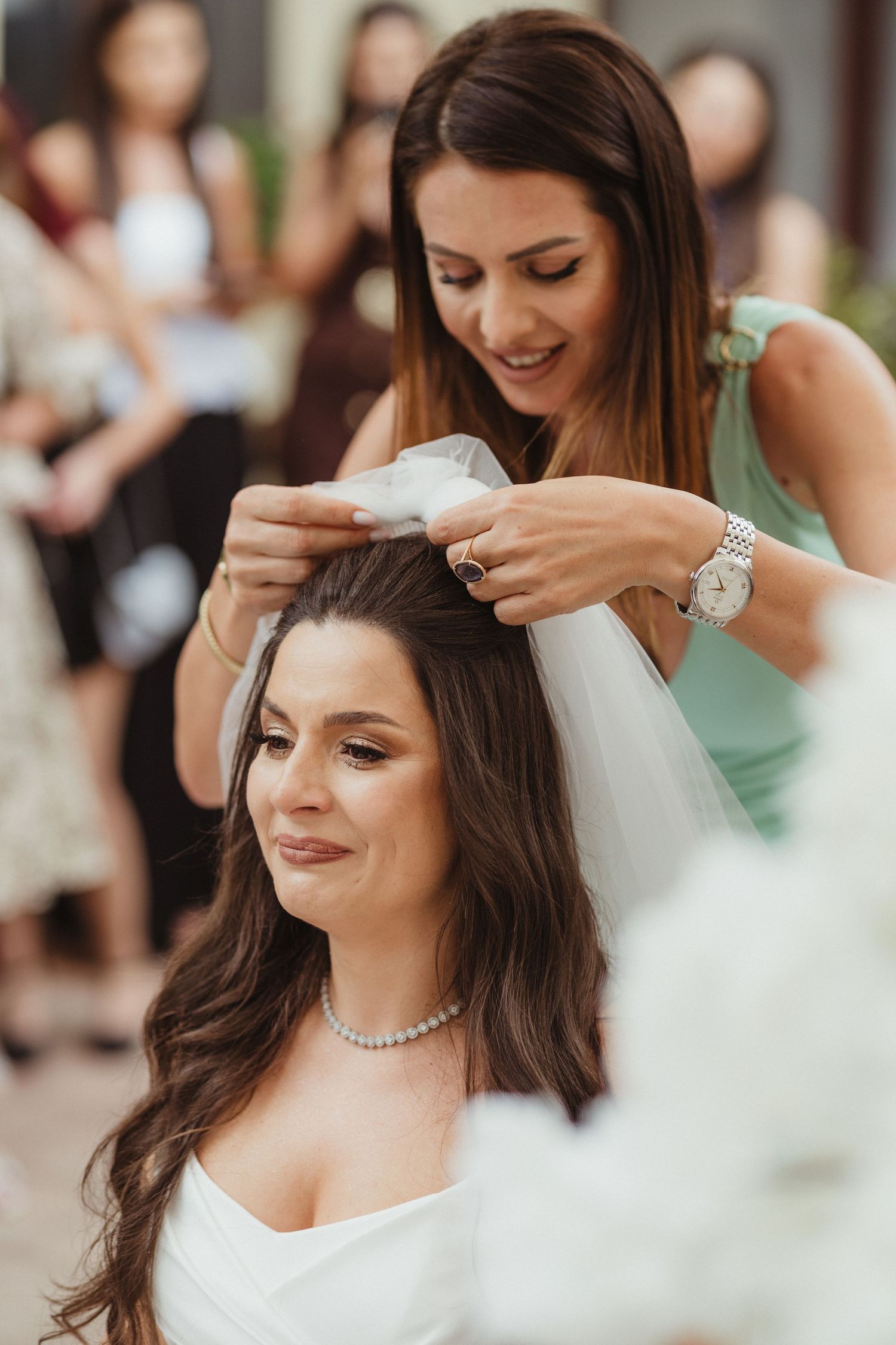 Bridesmaid placing white veil on brunette bride's head outdoors, guests visible in background.