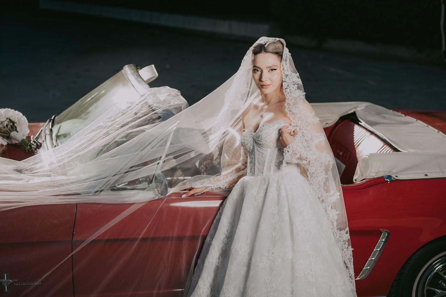 Elegant bride in a strapless ball gown with long veil poses beside a classic red convertible.