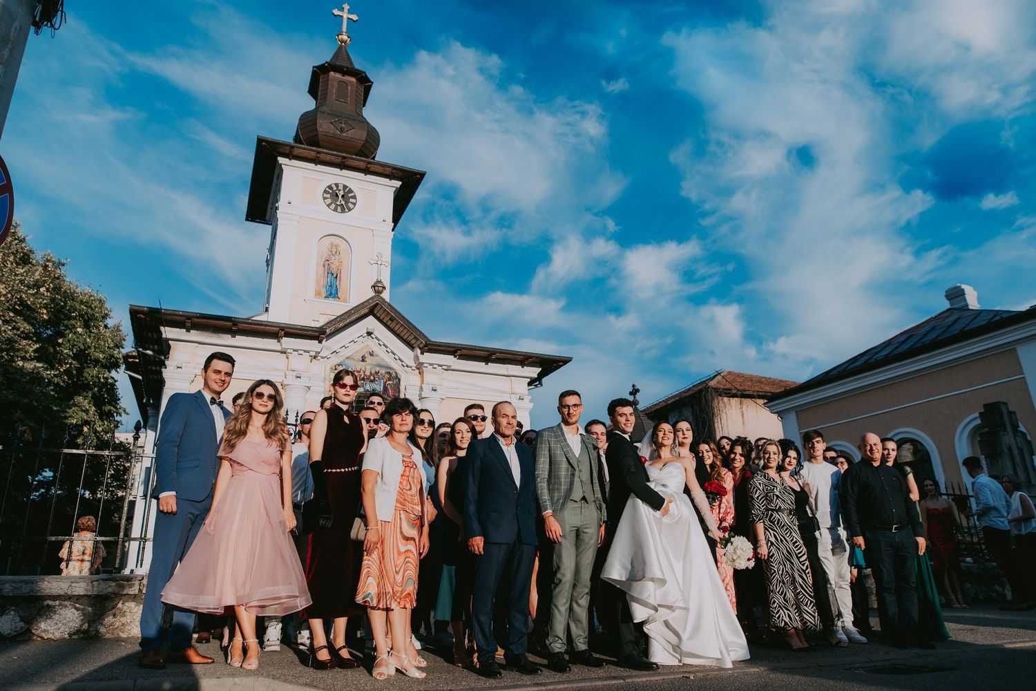 Wedding party group photo outside white Orthodox church under blue sky. Epicu Fotograf Drobeta-Turnu Severin
