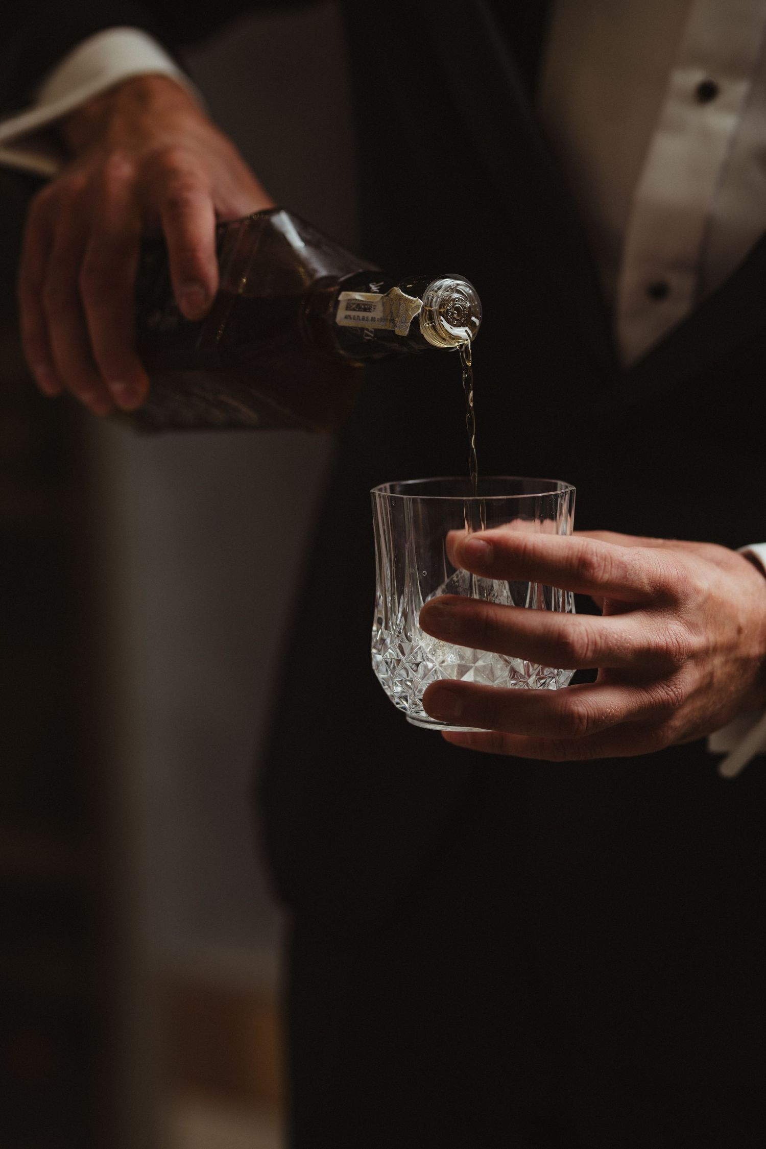 Groom in tuxedo pouring whiskey from bottle into crystal glass in dimly lit room.