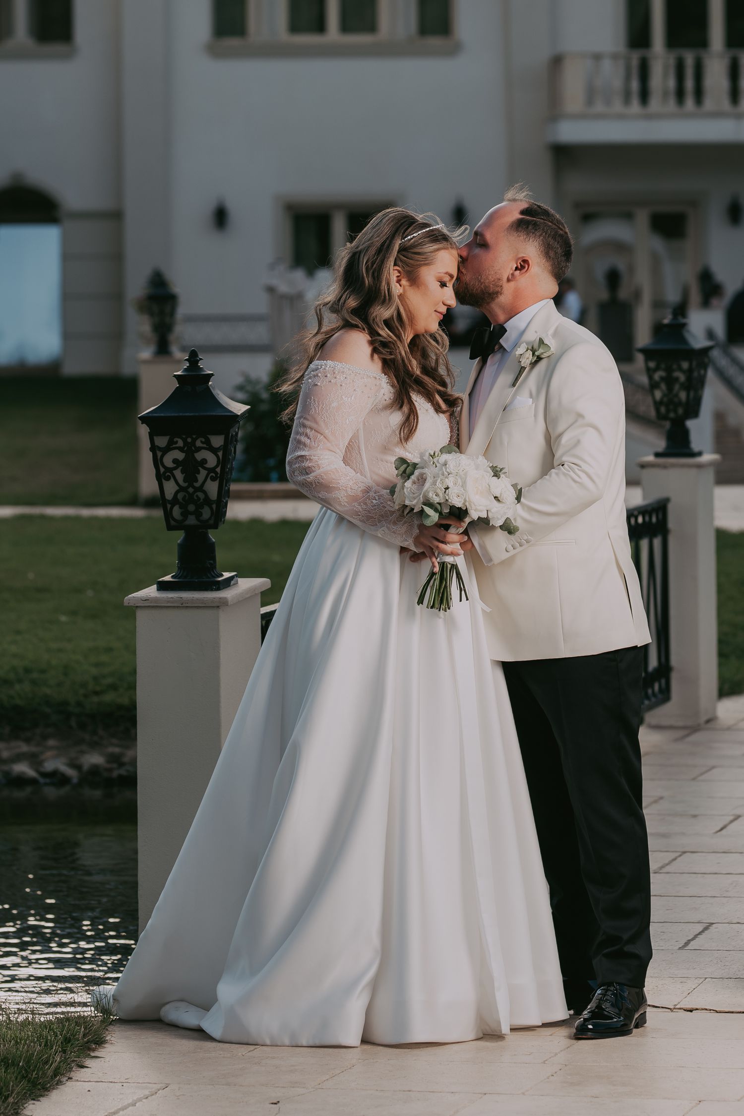 Bride and groom share tender kiss holding white floral bouquet beside a serene pond. Epicu Fotograf Drobeta-Turnu Severin