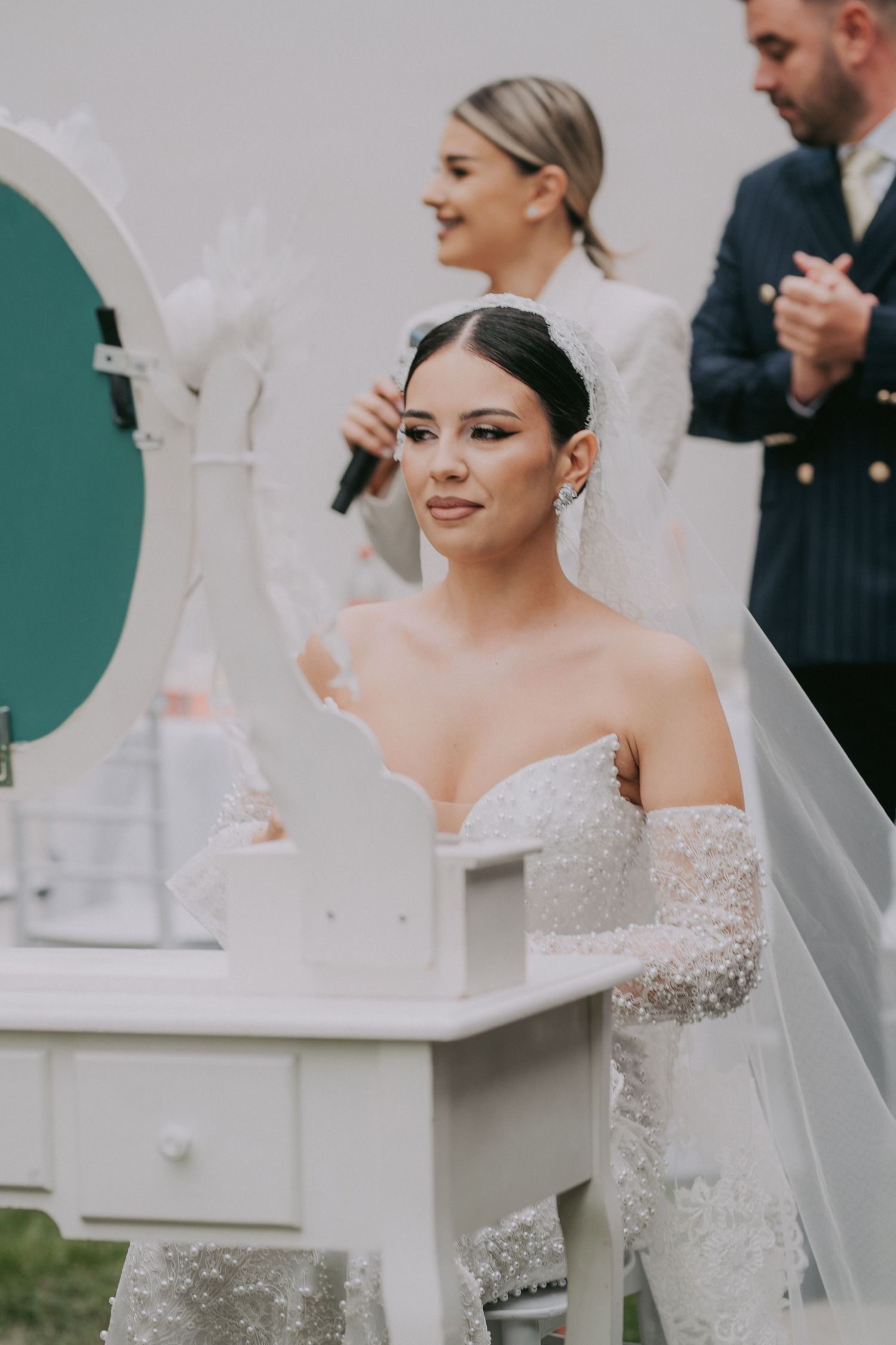 Beautiful bride in off-shoulder lace gown checking makeup at white vanity mirror during outdoor ceremony.