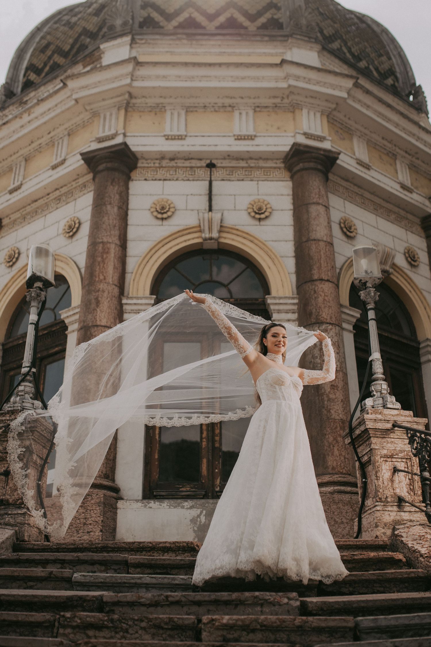 Bride in lace wedding dress lifts flowing veil dramatically before a grand neoclassical building.
