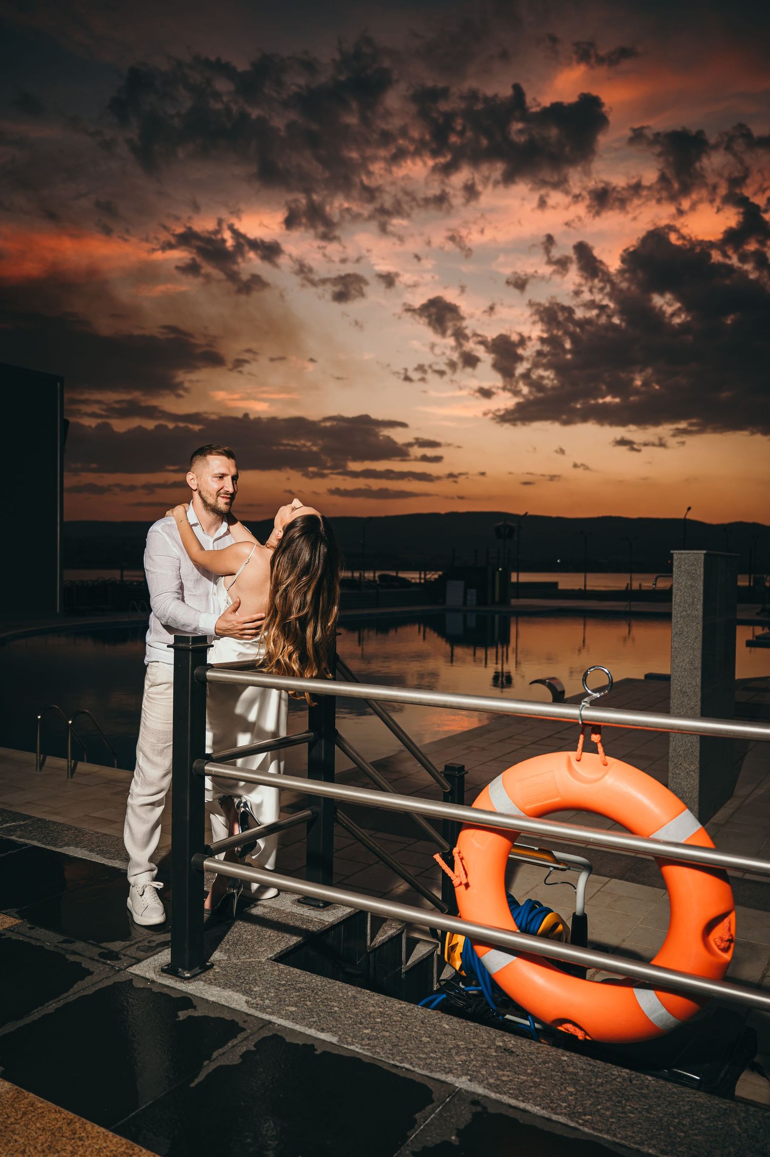 Romantic couple embracing on waterfront railing at dramatic sunset with orange life preserver nearby.