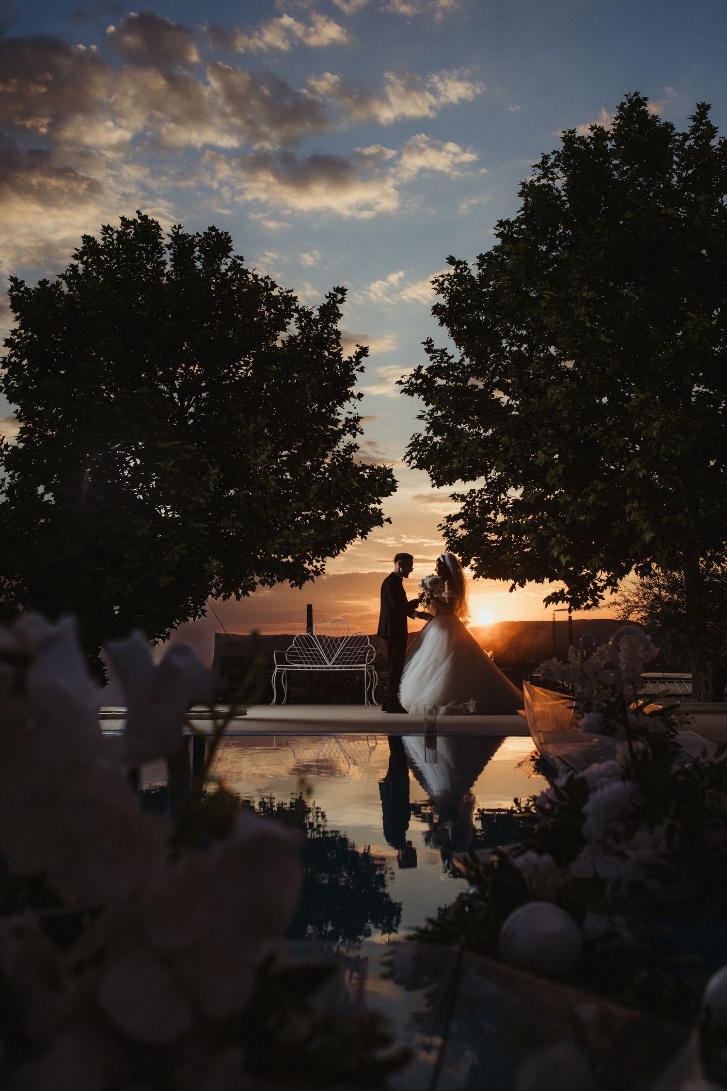 Silhouette of wedding couple holding hands by a reflective pool at golden sunset.