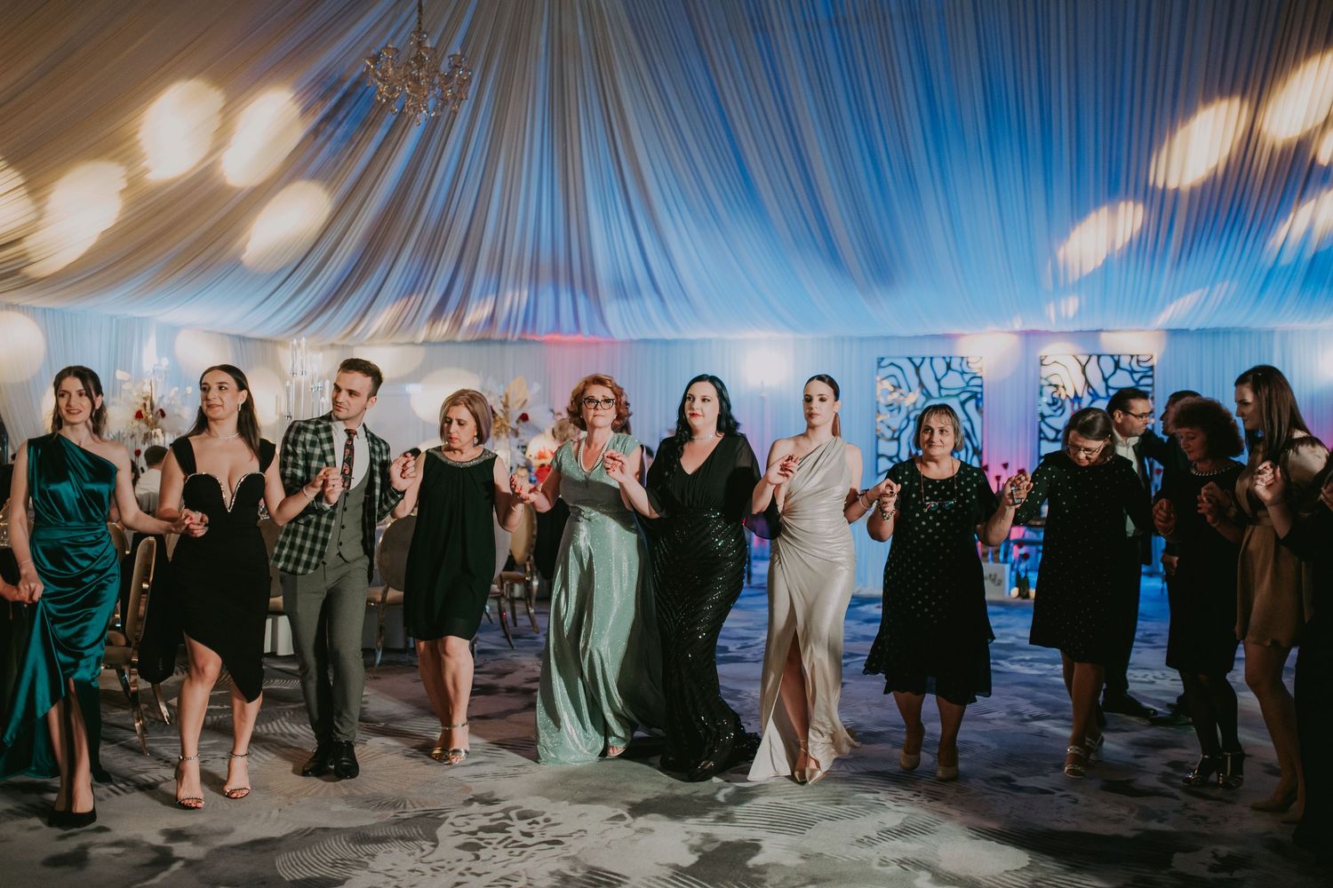Wedding guests in formal attire line up dancing together under blue lights in elegant tent venue. Epicu Fotograf Drobeta-Turnu Severin
