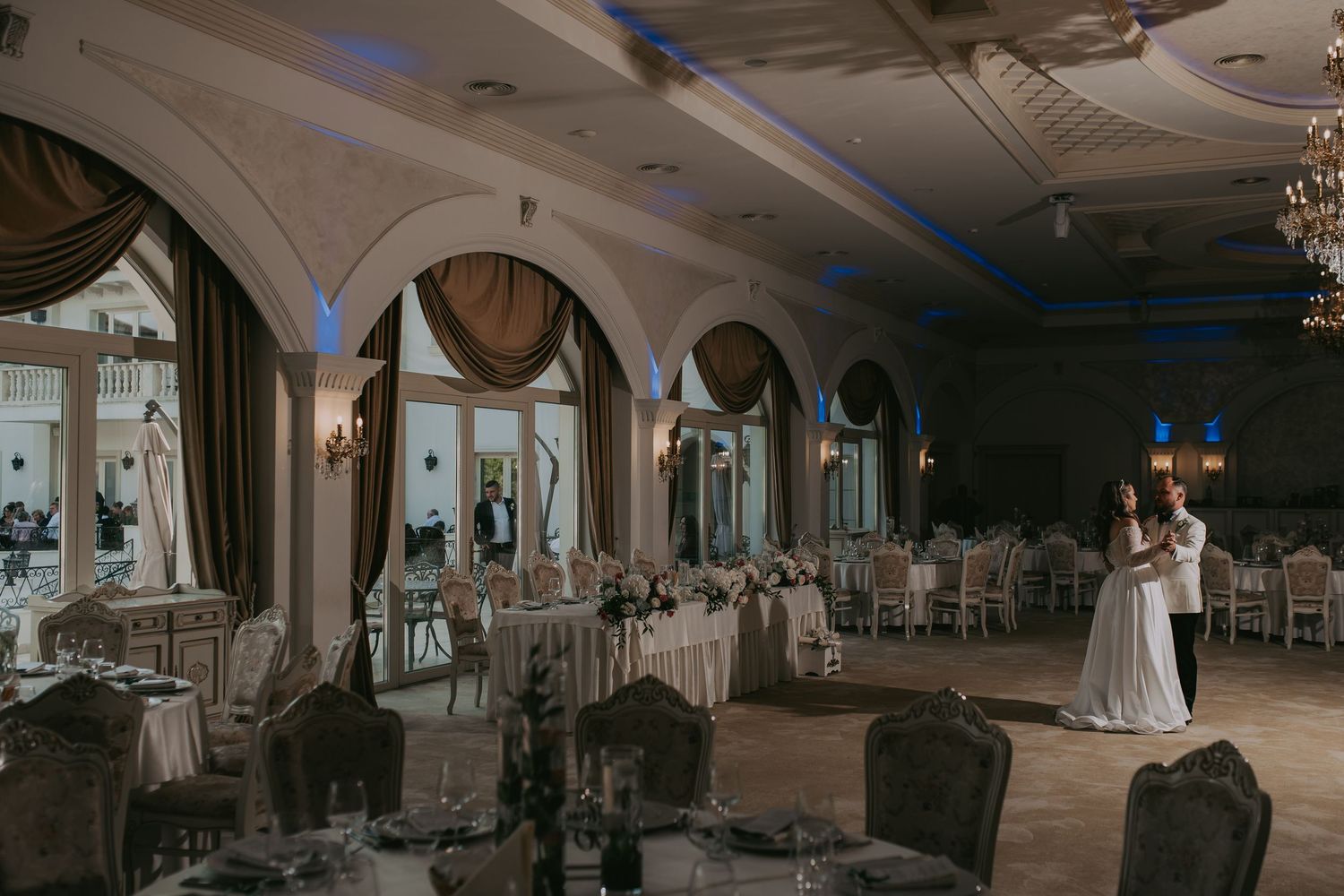 Bride and groom share first dance in elegant ballroom with arched windows and blue ambient lighting. Epicu Fotograf Drobeta-Turnu Severin