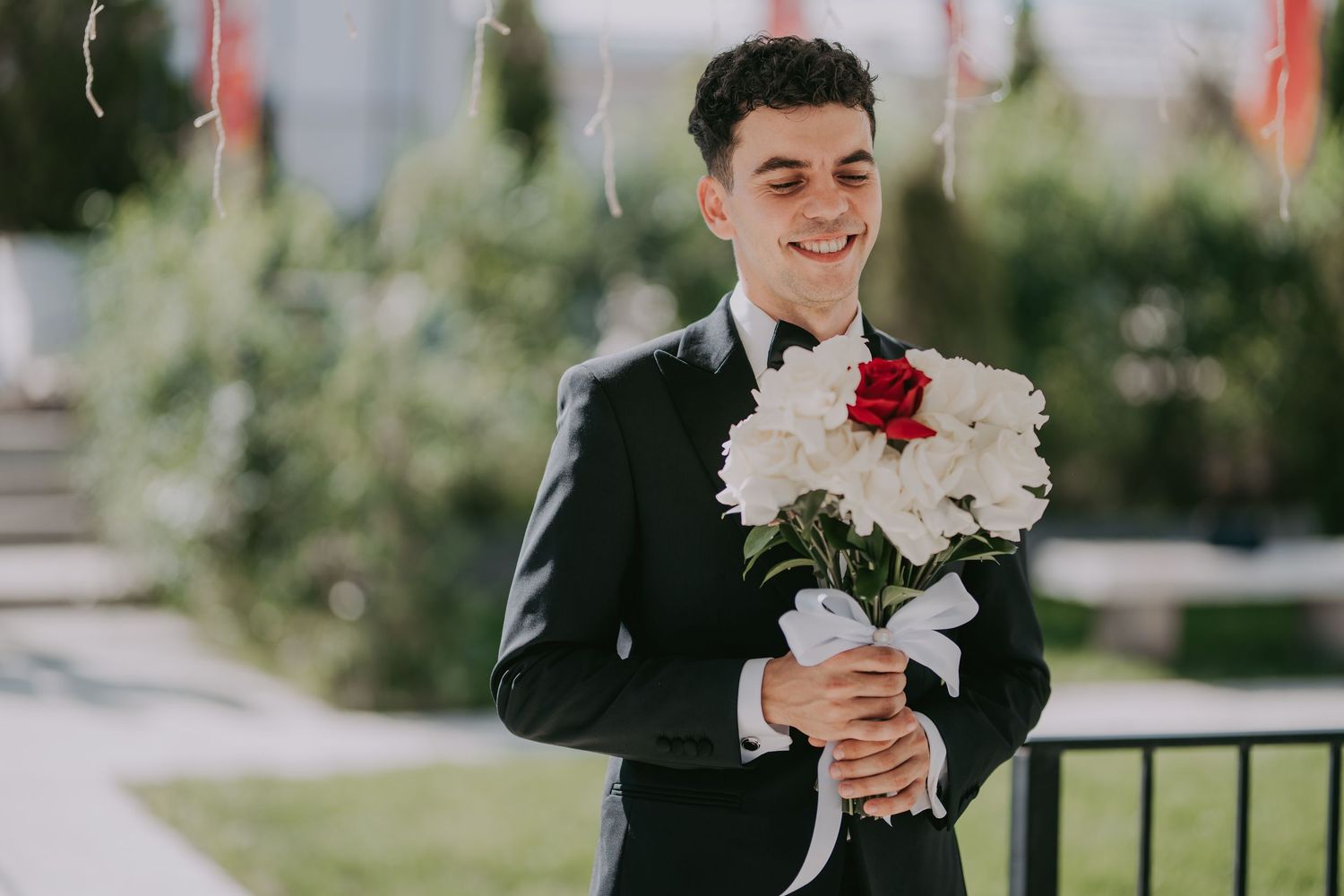 Smiling groom in black tuxedo holding a bouquet of white flowers with a red rose. Epicu Fotograf Drobeta-Turnu Severin