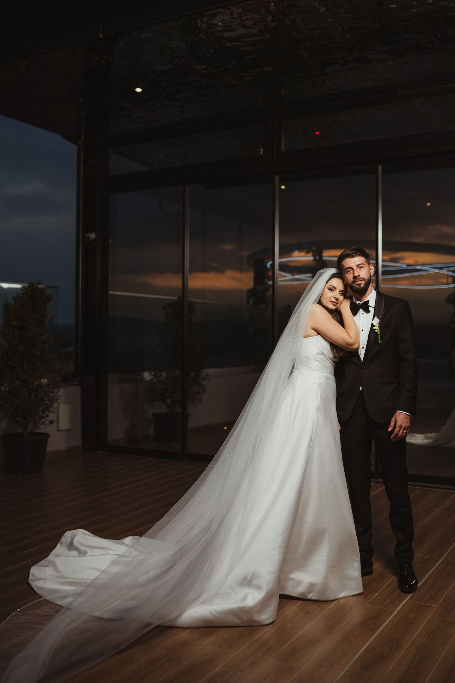 Bride in long white gown and groom in black tuxedo pose elegantly at dusk inside modern glass venue.