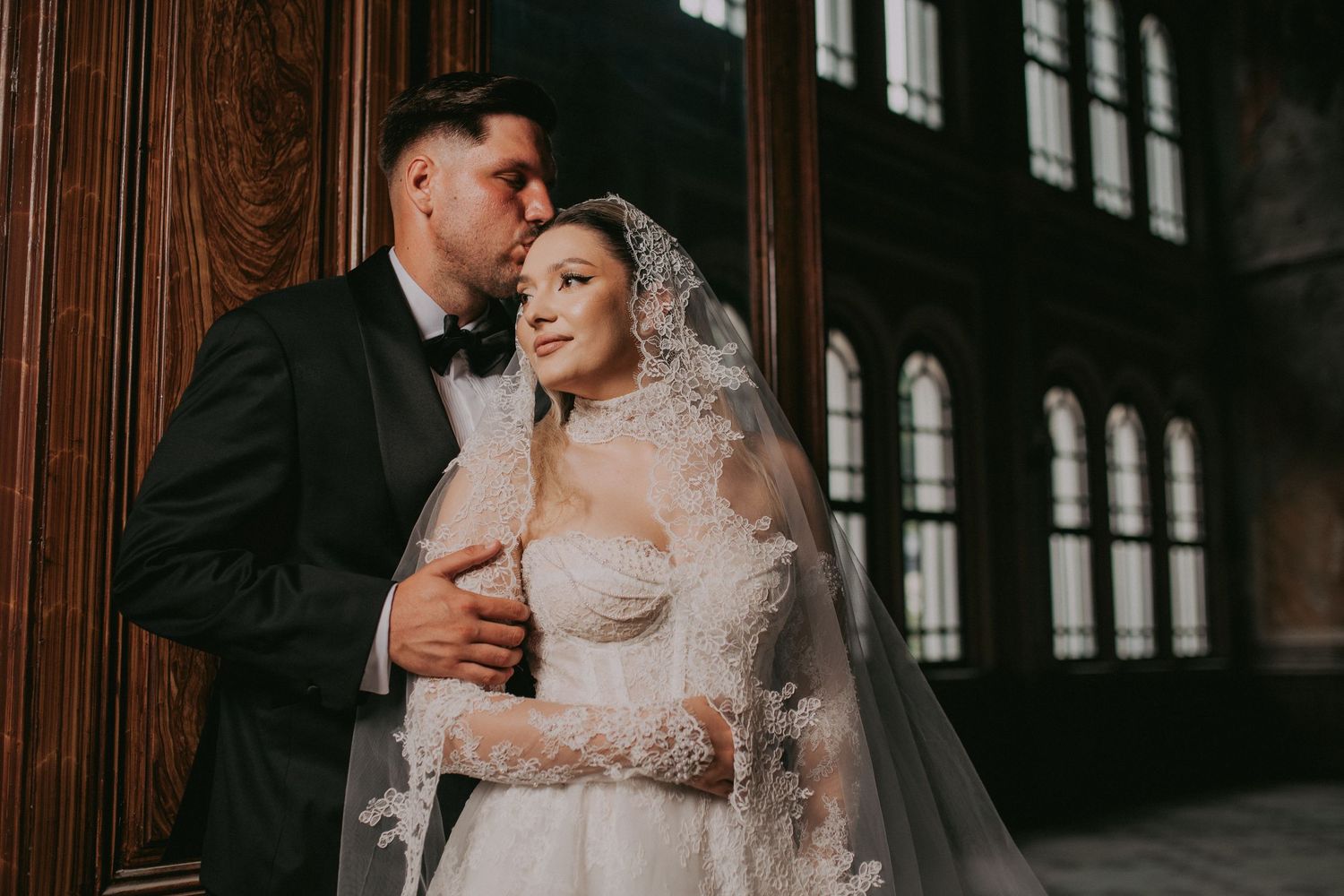 Groom in black tuxedo tenderly kissing bride in lace wedding dress and veil indoors.