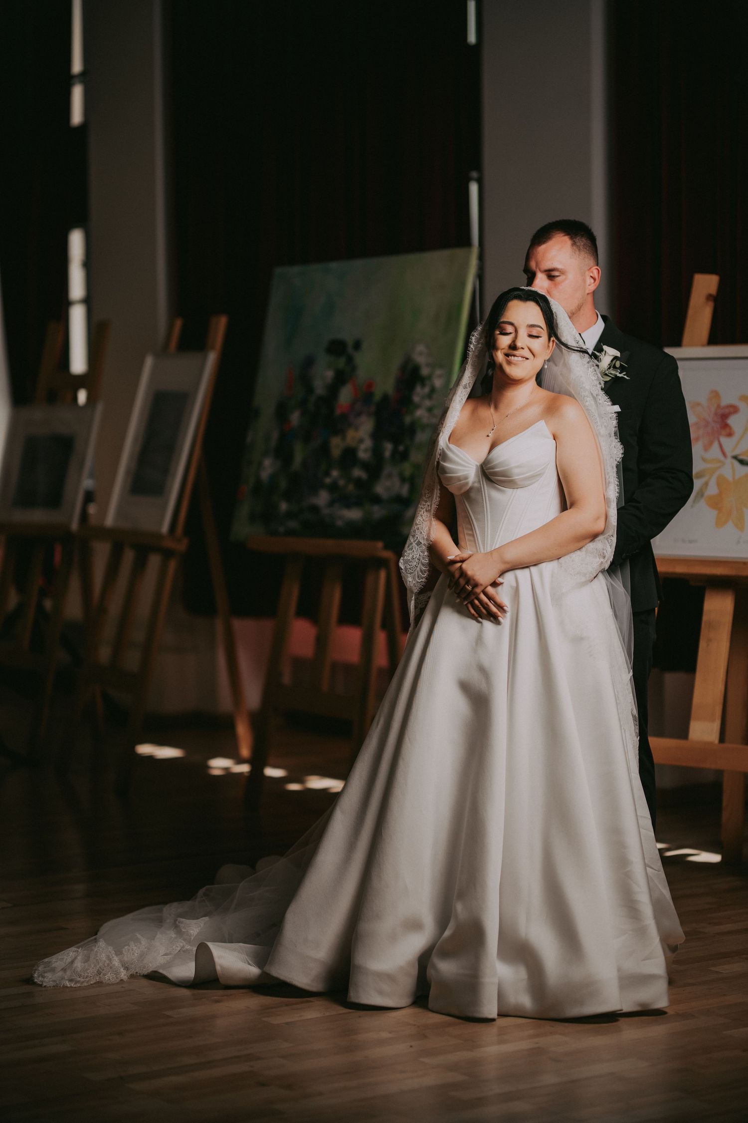 Elegant bride in white satin gown embraced by groom in art gallery with paintings on easels.