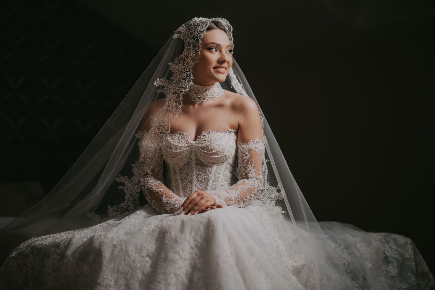 Elegant bride in a lace ballgown with ornate veil and headpiece posing in soft studio light.