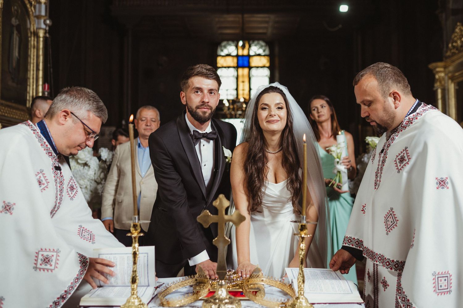 Bride and groom smile beside priests at Orthodox wedding altar with golden cross and ceremonial items.
