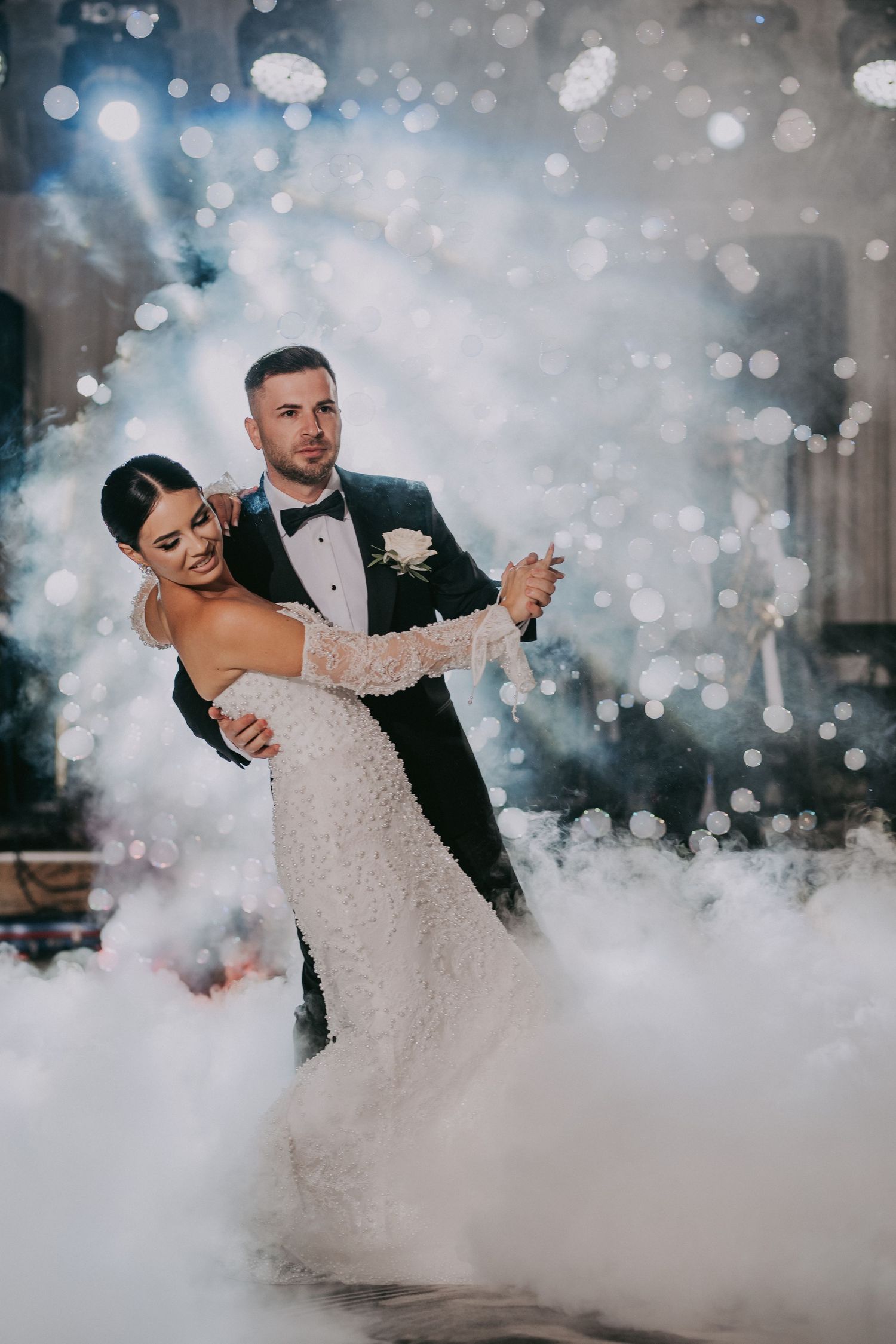 Bride and groom share their first dance surrounded by dramatic fog and sparkling bokeh lights at wedding reception.