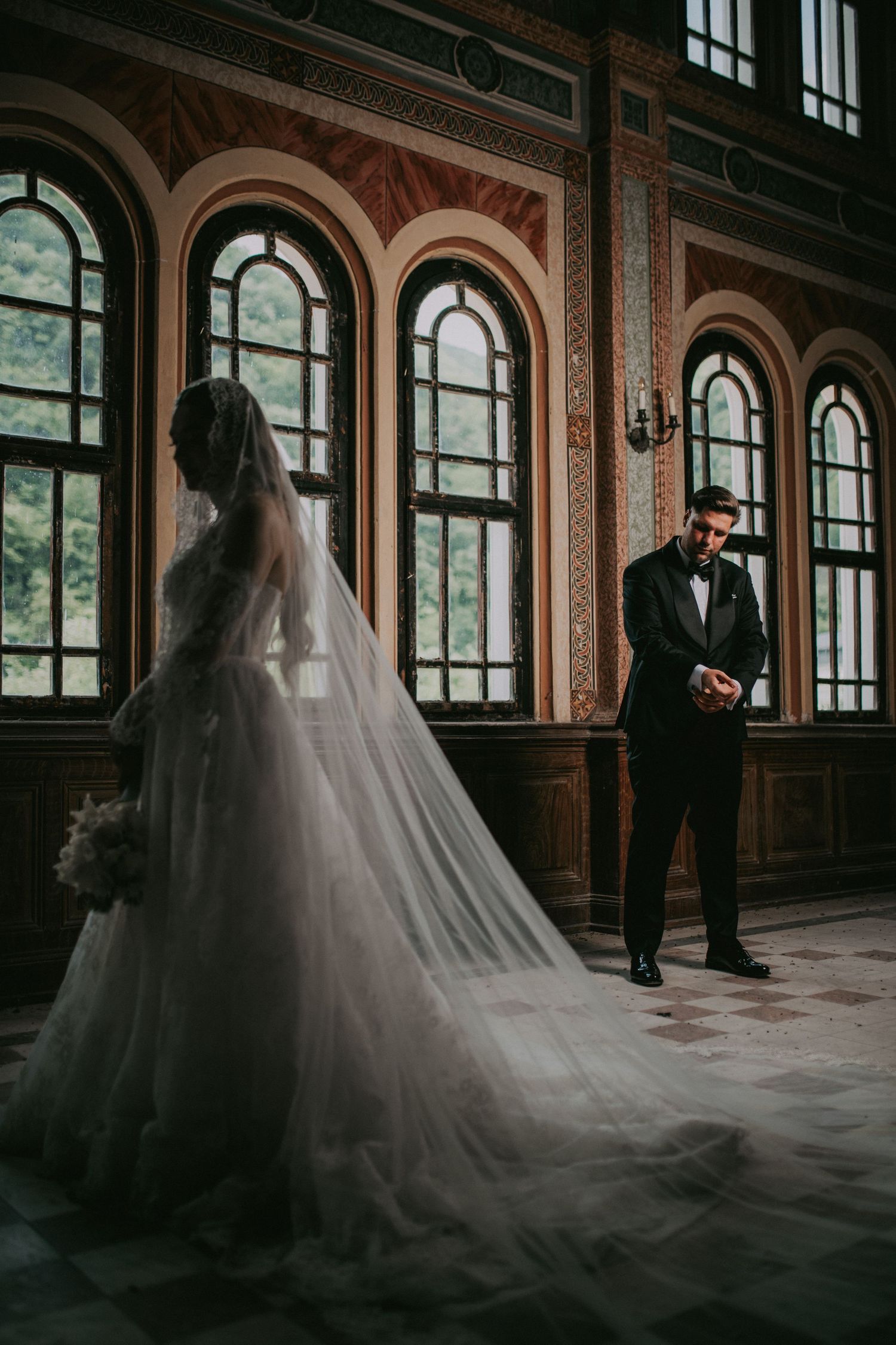 Groom emotional as bride in cathedral veil and ball gown approaches in ornate historic venue.