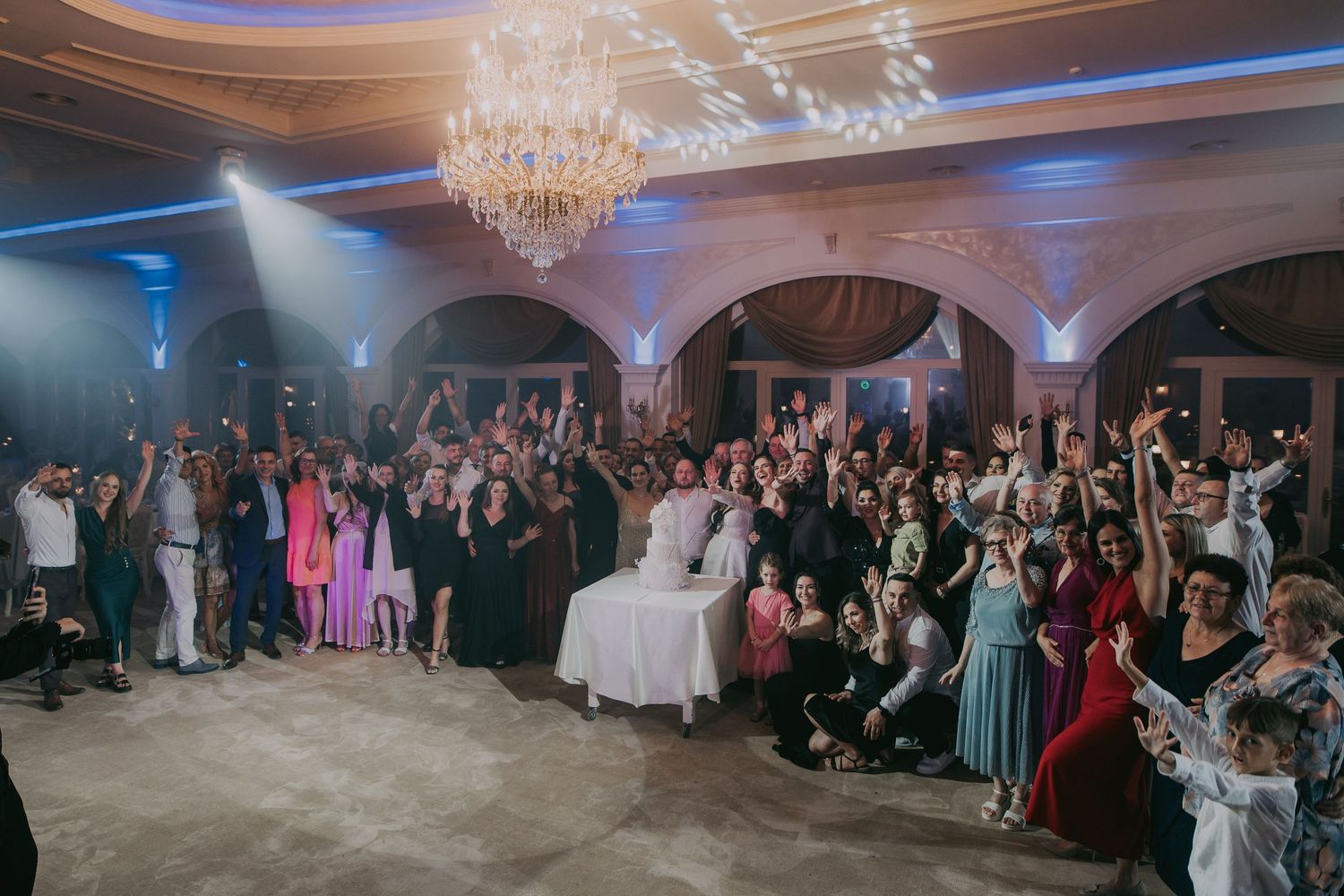 Large group of wedding guests celebrating together under chandeliers in an elegant ballroom venue. Epicu Fotograf Drobeta-Turnu Severin