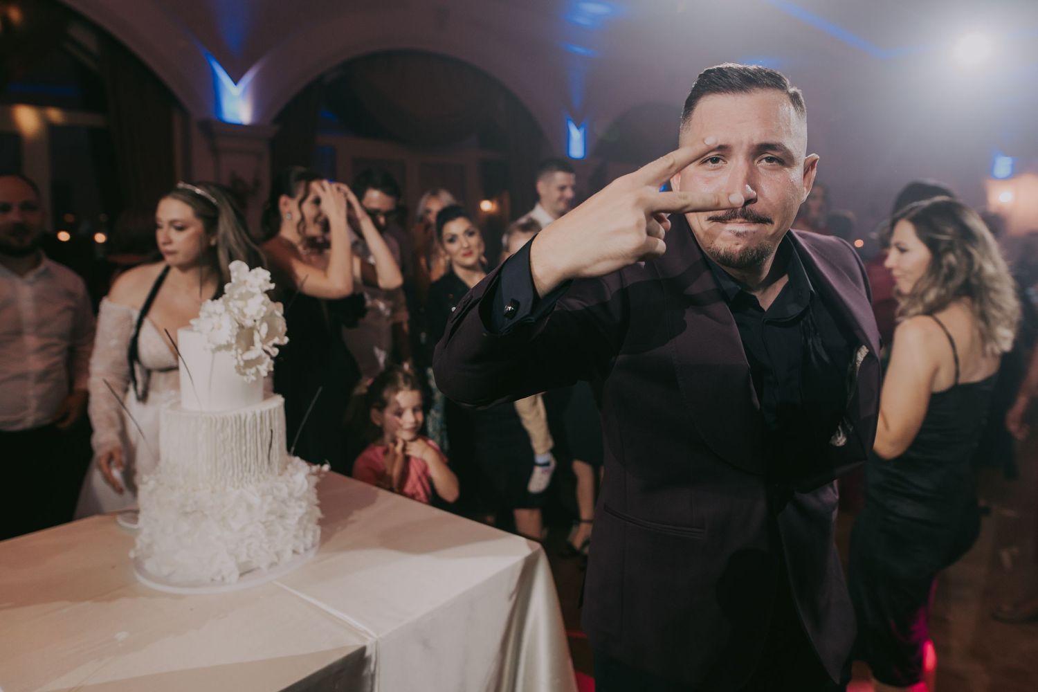 Man posing confidently beside a white wedding cake at a lively wedding reception celebration. Epicu Fotograf Drobeta-Turnu Severin