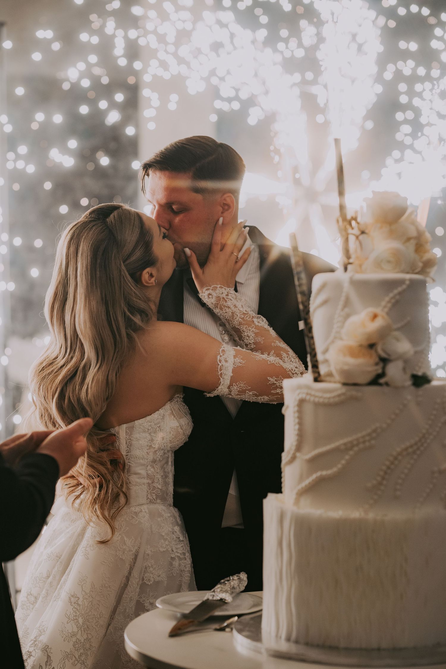 Bride and groom share passionate kiss while cutting elegant four-tier white wedding cake with roses.