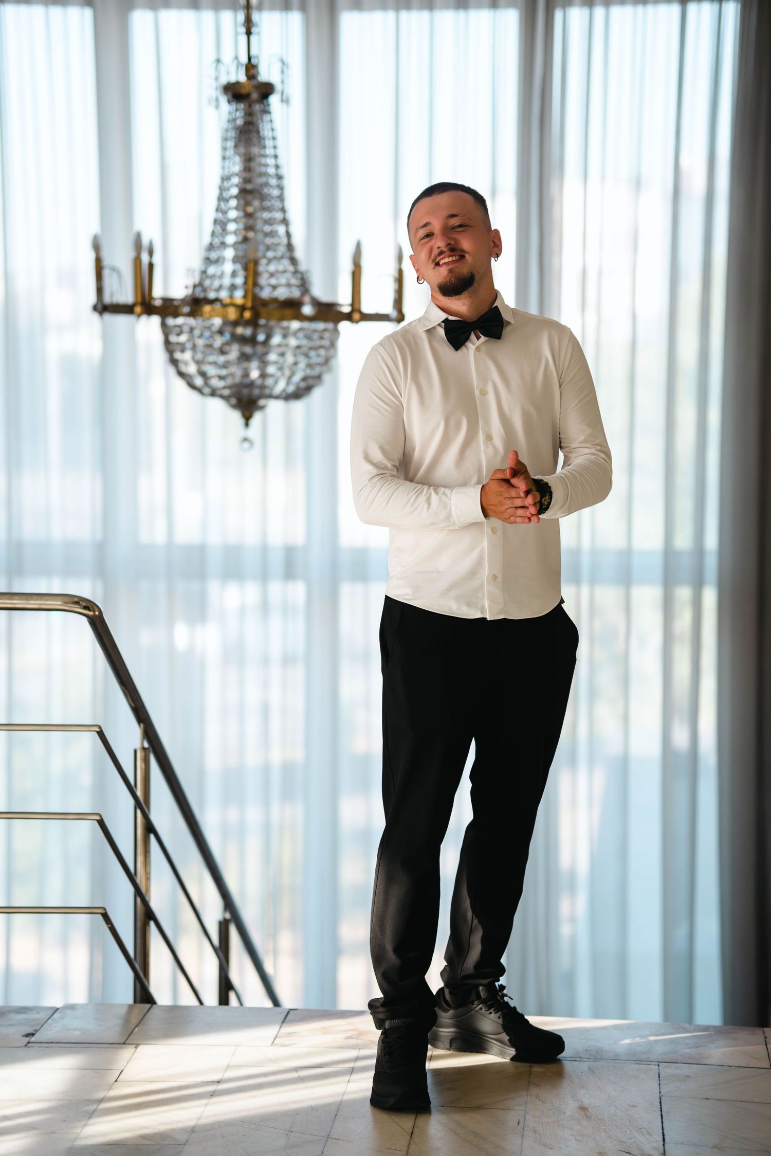 Smiling groom in white shirt and black bow tie standing near staircase under crystal chandelier indoors.Epicu Fotograf Drobeta-Turnu Severin