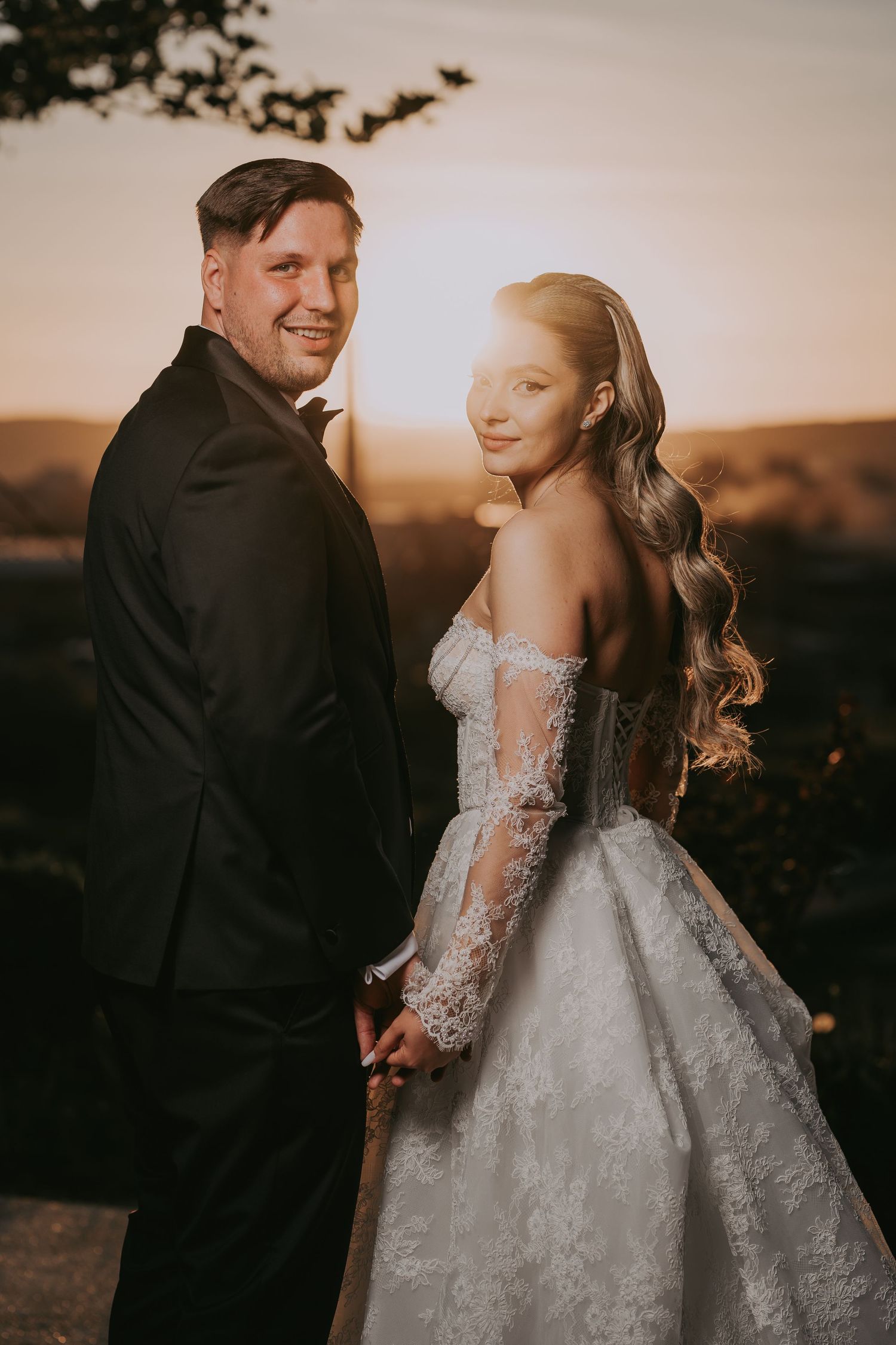 Bride and groom holding hands at golden sunset, she wears off-shoulder lace gown with long sleeves.
