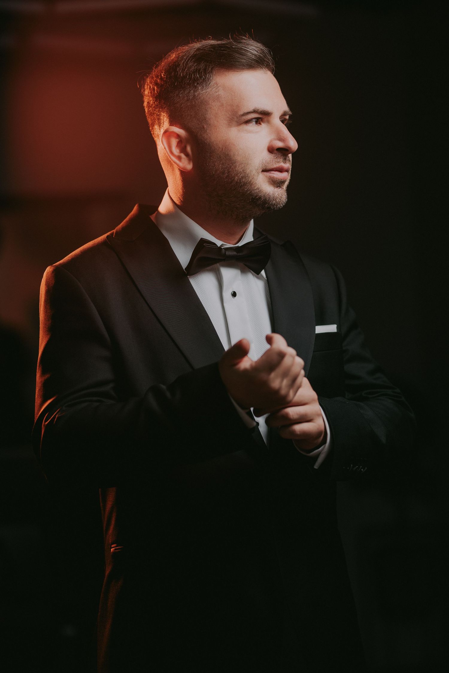 Dapper groom in black tuxedo with bow tie adjusting cufflinks in warm moody dark background lighting.