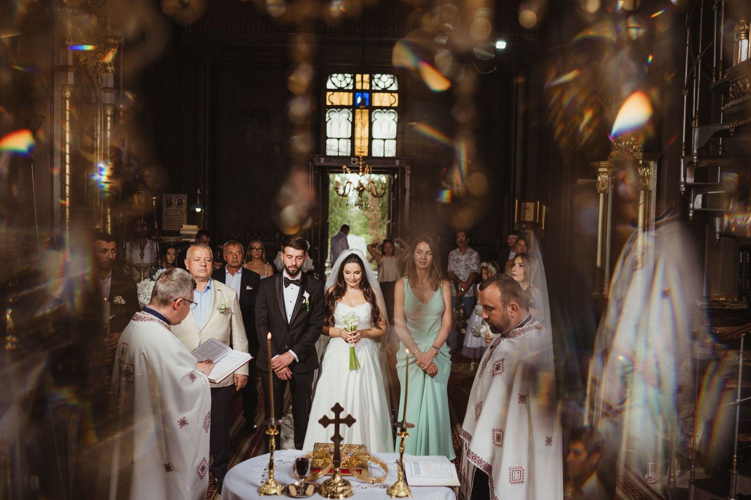 Bride and groom stand at the altar during an Orthodox church wedding ceremony with golden candlesticks and stained glass.