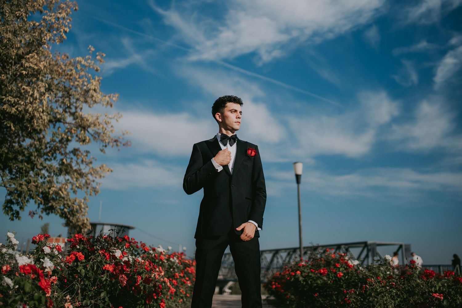 Dapper groom in black tuxedo with red rose boutonniere adjusting bow tie outdoors against blue sky. Epicu Fotograf Drobeta-Turnu Severin
