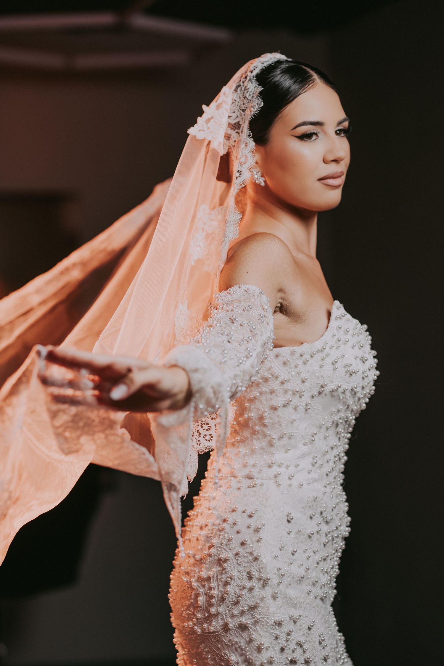 Bride in beaded mermaid wedding dress extends arm with veil flowing in light.