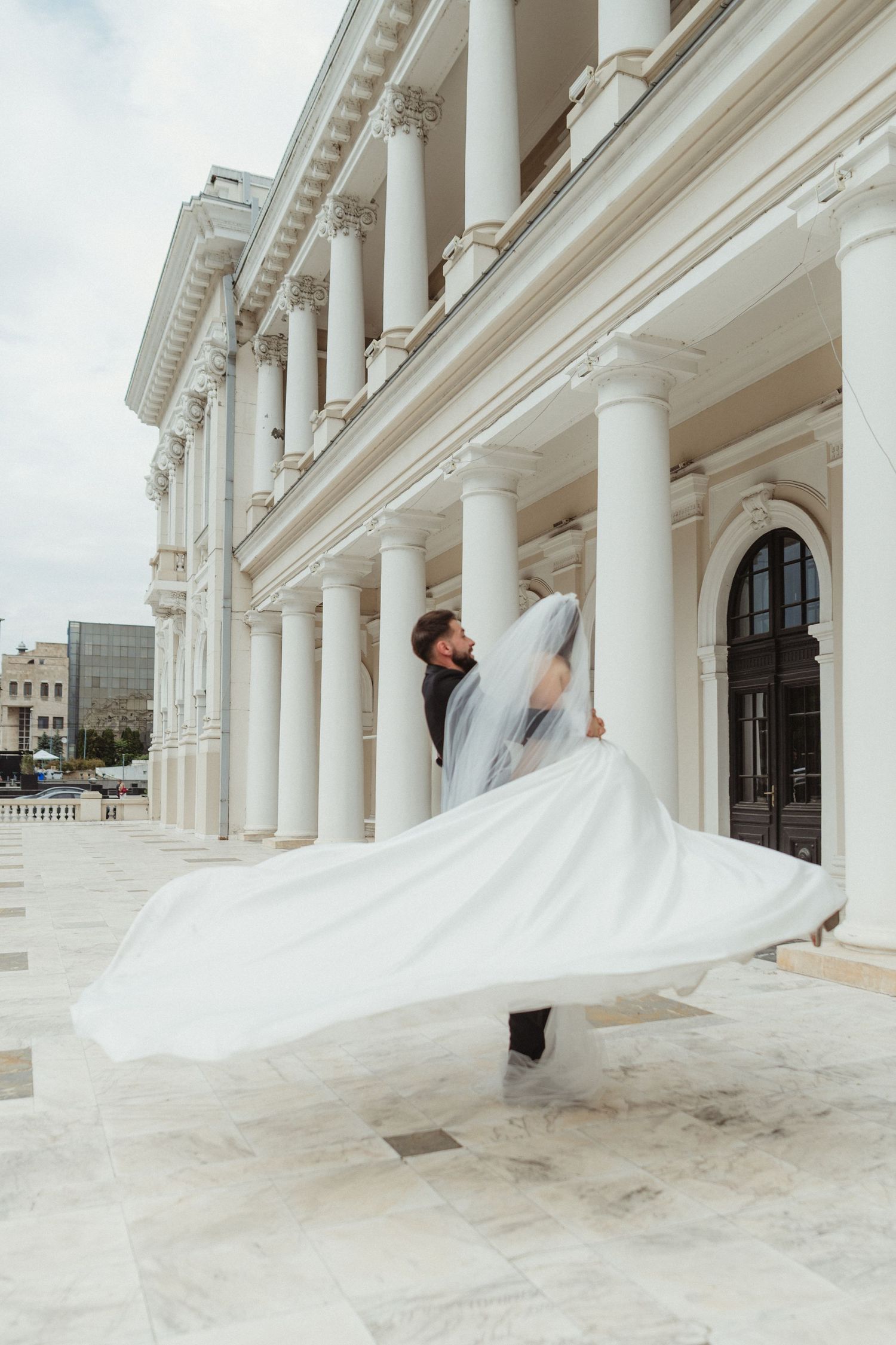 Groom spinning bride in a flowing white wedding gown outside a neoclassical building with columns.