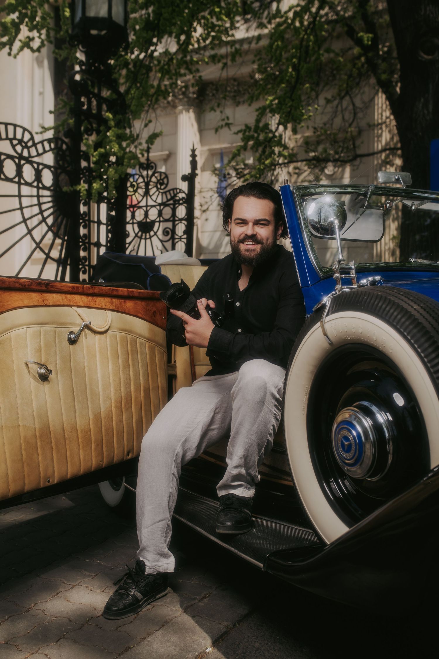 Smiling man with beard sitting on vintage classic car holding camera in outdoor urban setting.