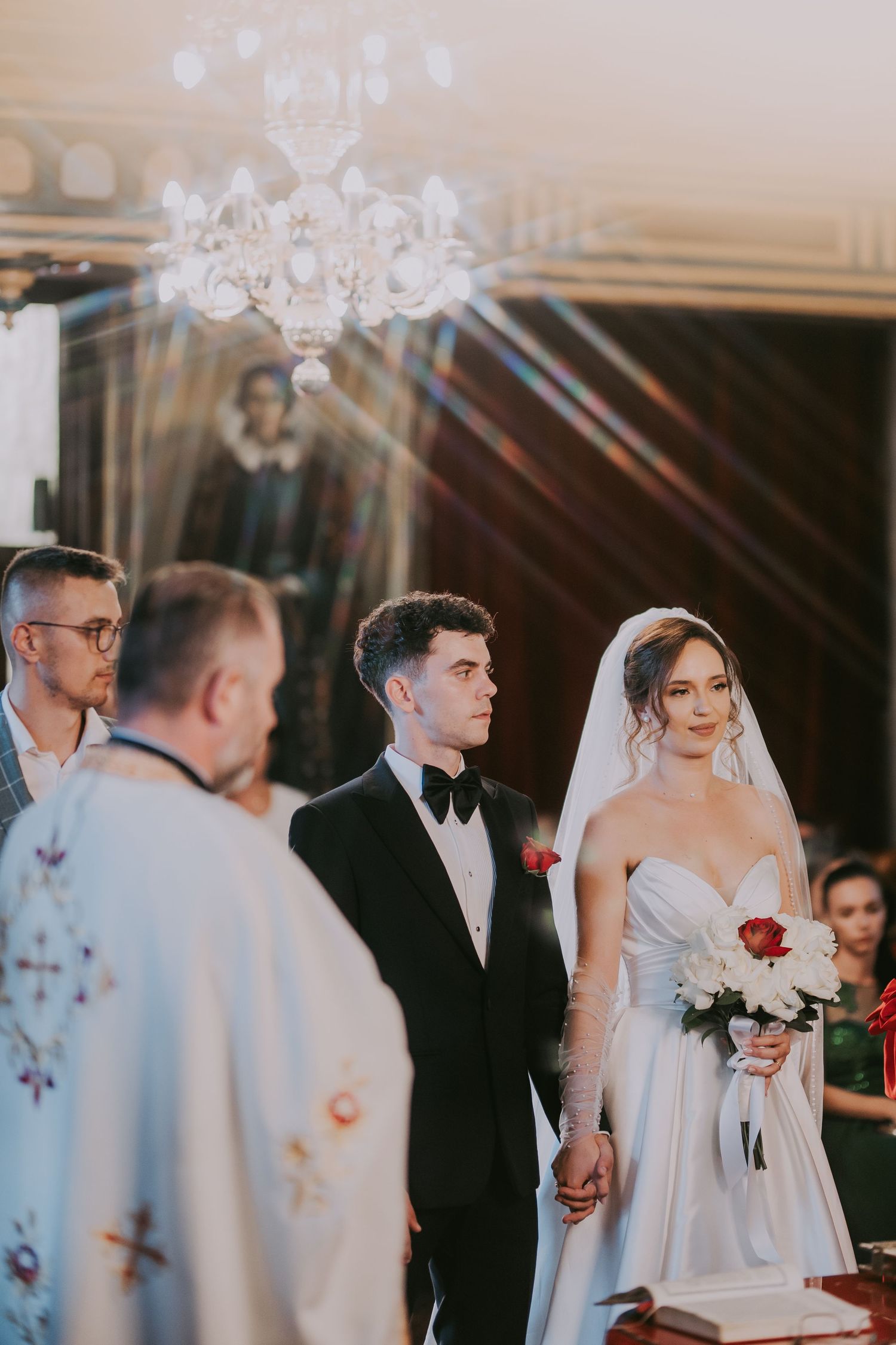 Bride and groom hold hands before Orthodox priest during church wedding ceremony with chandelier above. Epicu Fotograf Drobeta-Turnu Severin