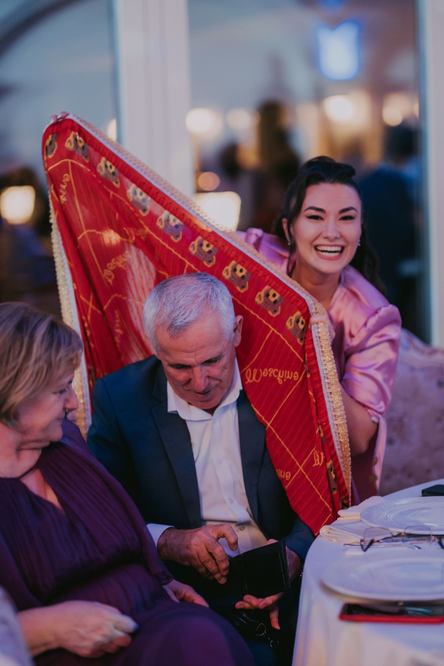 Smiling Asian bride in pink dress playfully hiding behind a red embroidered blanket at wedding reception. Epicu Fotograf Drobeta-Turnu Sever