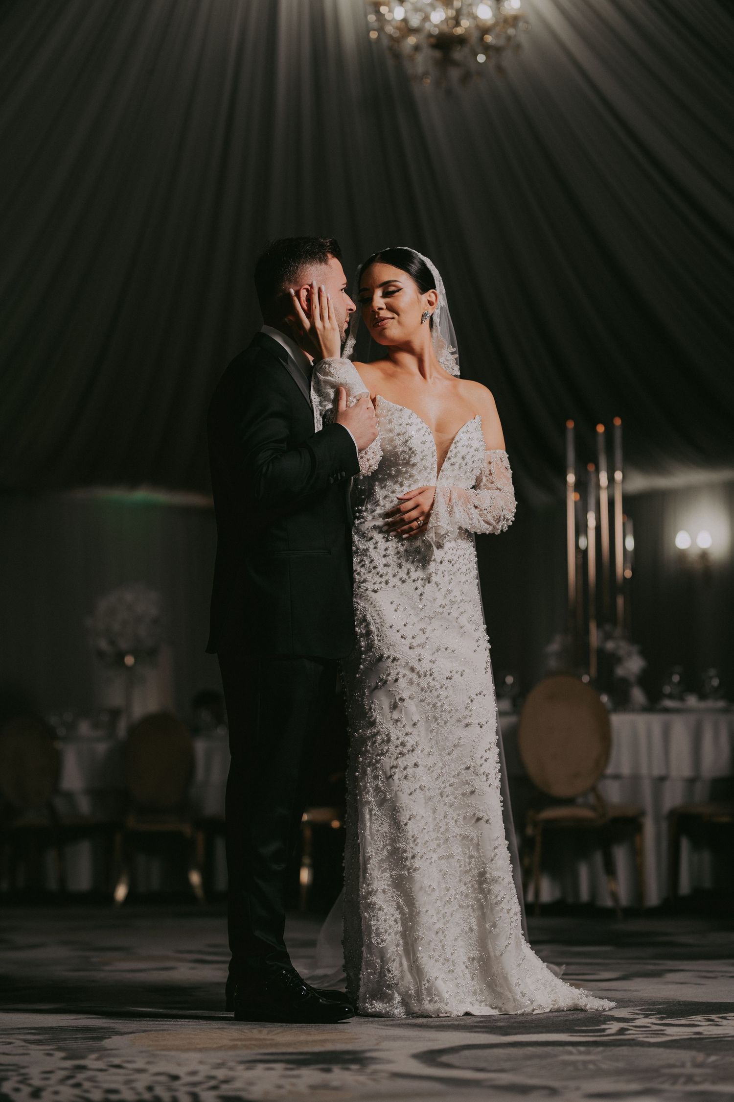 Emotional groom covers face beside smiling bride in embellished wedding gown at reception.